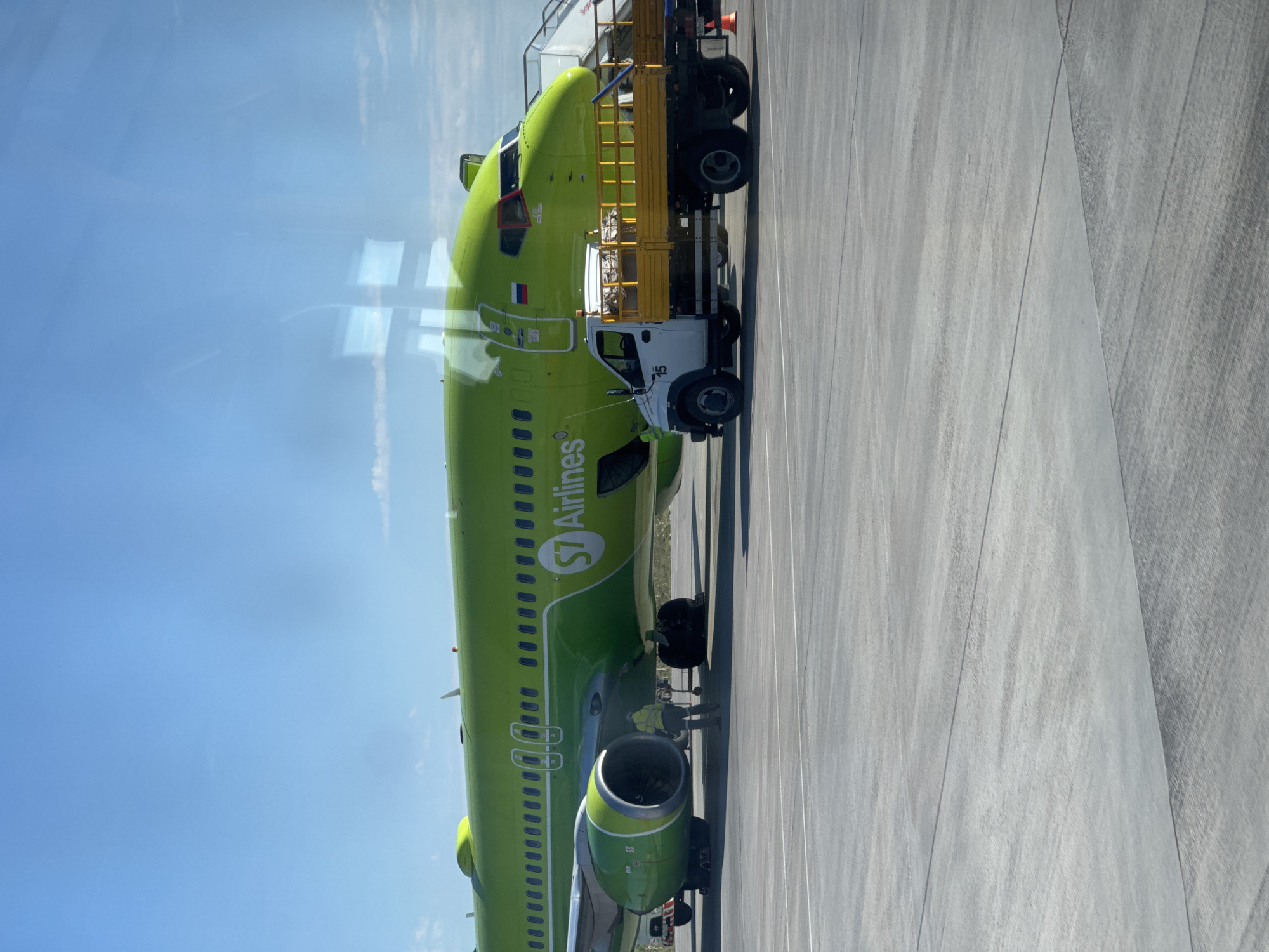 An S7 Airlines airplane is parked on the tarmac at Chulman Airport (UUDJ) in Chulman, Russia. The aircraft is predominantly green with the S7 Airlines logo on its fuselage. A small white utility truck with a yellow flatbed is parked next to the airplane. Two individuals, one wearing a yellow high-visibility vest, are standing near the plane's engines. The sky is clear blue with a few wispy clouds. The ground is paved with concrete.