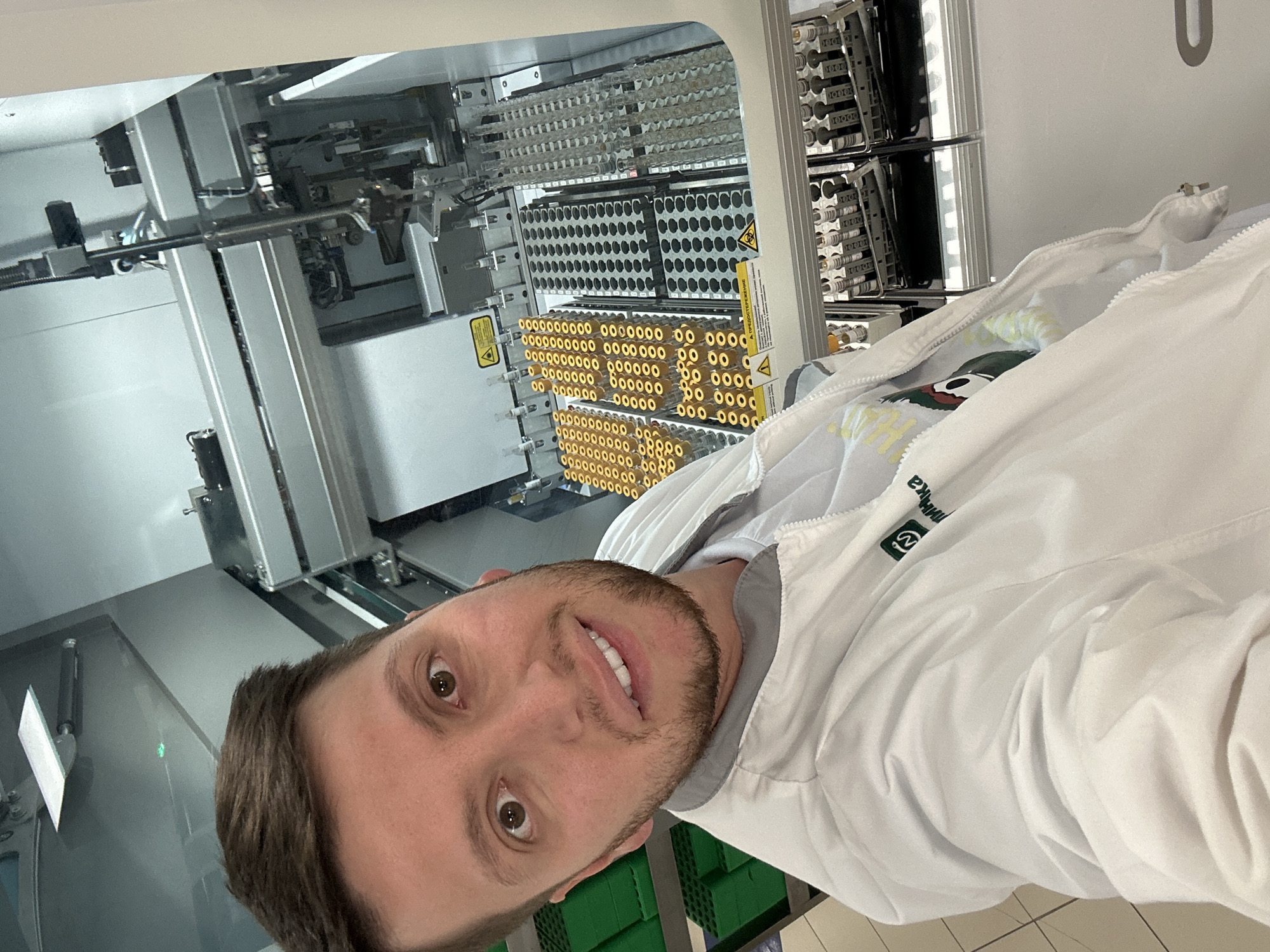 A young adult male, of light complexion with short brown hair and a neatly trimmed beard, is centered in a selfie taken in what appears to be an automated medical laboratory in Moscow, Russia. He is smiling directly at the camera. He wears a white lab coat with a grey collar, unzipped to reveal a white t-shirt underneath featuring a colorful graphic. On the right chest area of his lab coat (viewer's left), a green logo is visible, containing a stylized symbol and the Cyrillic word "КЛИНИКА" (Klinika, meaning Clinic).
Behind the man, a large, modern automated laboratory analyzer occupies most of the background. Inside the transparent cover of the machine, numerous racks are filled with various biological sample containers. Many racks hold clear glass or plastic vials with bright yellow caps, arranged in neat rows. Other sections contain racks with clear, uncapped tubes or tubes with clear stoppers, suggesting different types of samples. A robotic arm with a pipetting mechanism is visible, positioned over some of the sample trays, indicating automated processing. Two yellow triangular warning signs are prominently displayed within the machine's interior; one clearly shows the Russian word "ПРЕДОСТЕРЕЖЕНИЕ" (Predosterezhenie, meaning CAUTION or WARNING), while the other bears a universal biohazard symbol. To the left, behind the man's shoulder, several green plastic racks or containers are stacked on a metal shelving unit. The floor is covered with clean white tiles.