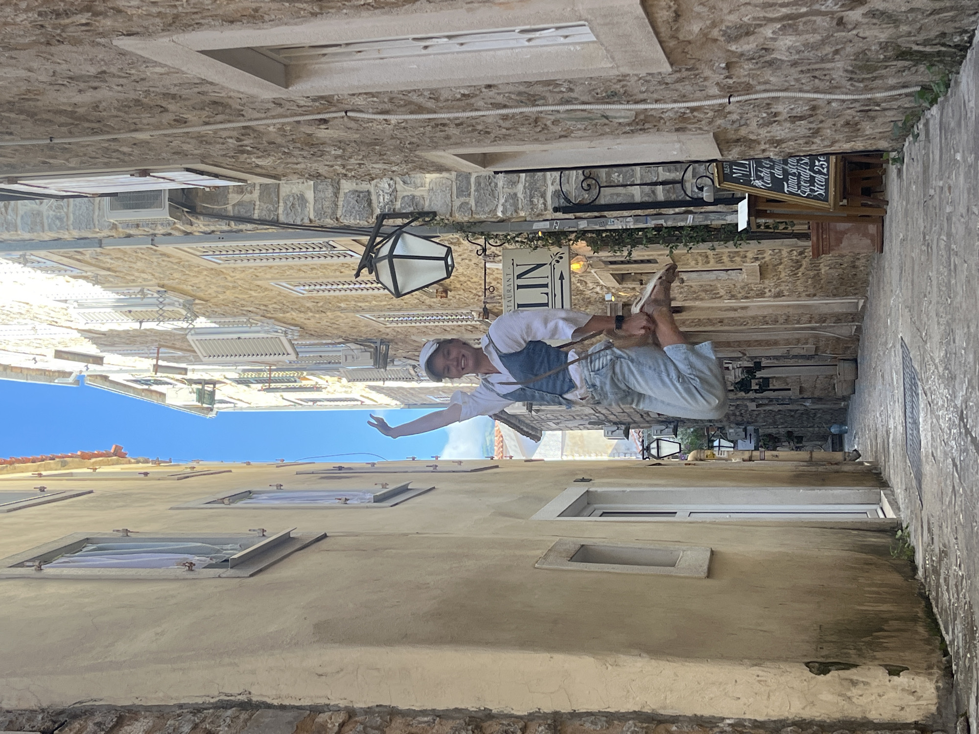 A young man is captured mid-pose, appearing to levitate horizontally between old stone buildings in a narrow alleyway. He wears a white t-shirt, light blue overalls or vest, and light-colored trousers, along with a cap. He is smiling brightly, with his right arm extended straight out and his left arm bent, creating the illusion of defying gravity.
The setting is a picturesque, stone-paved street, characteristic of an old town, likely in Budva, Montenegro. The buildings are constructed from stone and some have a light yellowish-beige facade. Numerous windows, some with white shutters or modern ventilation units, dot the walls. Overhead, a clear, vibrant blue sky is visible, indicating it's daytime with good weather. An ornate black lantern hangs from one building, and greenery trails from some ledges.
Visible text on a sign above the person's head reads "RESTAURANT LIN" with an arrow pointing right. Below it, a blackboard menu advertises "Catch of day," "Tuna stea" (likely Tuna steak), "Swordfish," and "Steak 25€," indicating a local eatery. The overall scene suggests a cheerful and playful moment within a historic European street.