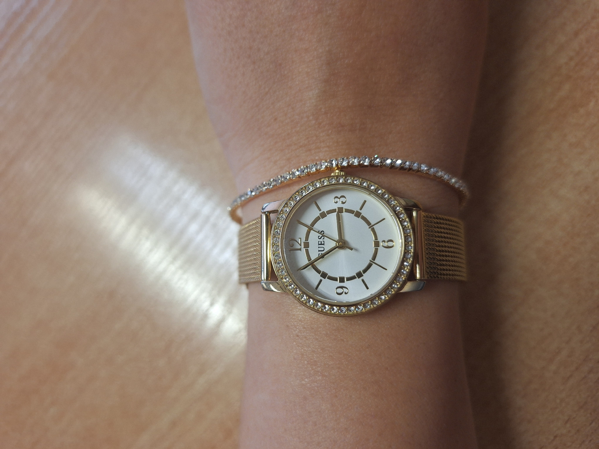 A close-up, high-angle shot captures a person's left arm, adorned with a gold-toned wristwatch and a sparkling bracelet, resting on a light brown surface, possibly a wooden table or desk. The setting is indoors, likely in Varna, Bulgaria.
The main subject is a round Guess brand wristwatch, featuring a white or cream dial with minimalist hour markers and prominent numbers at 12, 3, 6, and 9. The watch hands indicate a time of approximately 10:20 or 10:21. The watch case and mesh strap are gold-toned, and its bezel is encrusted with a single row of small, clear stones or crystals. Below the 12 o'clock marker, the brand name "GUESS" is clearly visible in gold lettering on the dial.
Just above the watch, a delicate gold-toned bracelet encircles the wrist. This bracelet is thin and flexible, entirely encrusted with numerous small, clear, brilliant-cut stones, resembling a tennis bracelet.
The skin of the arm appears light to medium-toned, with visible pores. The background surface exhibits subtle grain patterns and reflections of light, creating an impression of texture and depth. The lighting is soft but highlights the metallic and crystalline details of the jewelry.