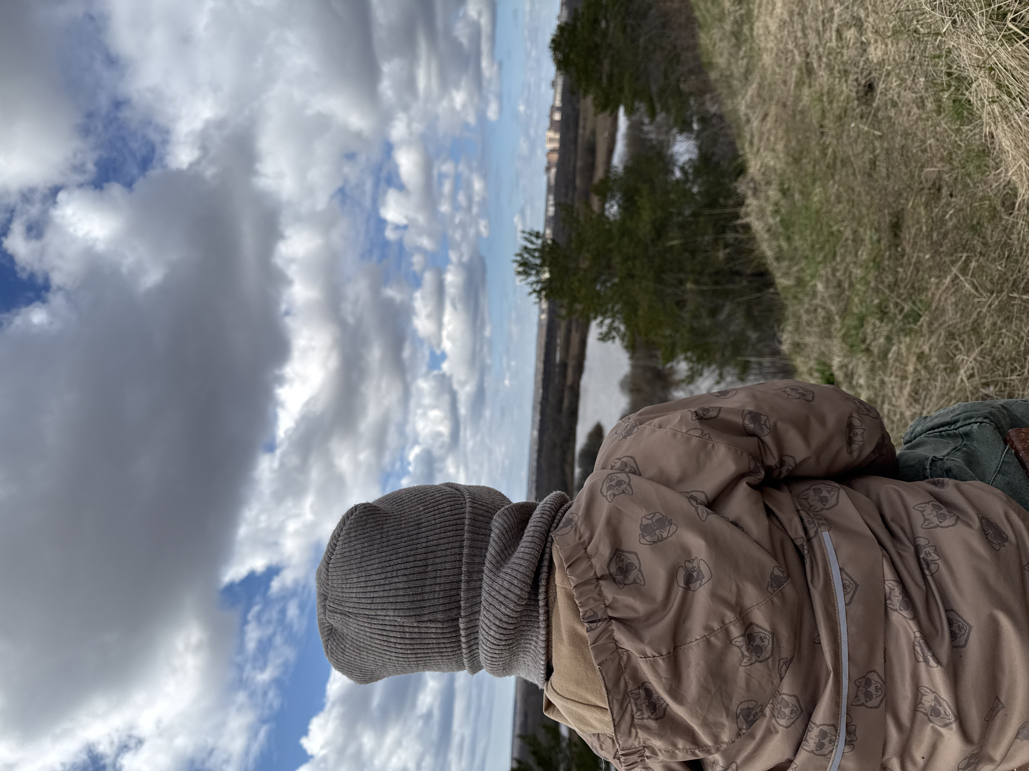 A person wearing a gray knit hat and a patterned brown jacket sits on a grassy hill overlooking a river or lake. In the background, buildings are visible on the opposite shore, and the sky is filled with cumulus clouds. The photograph may be taken in Glazov, Russia.