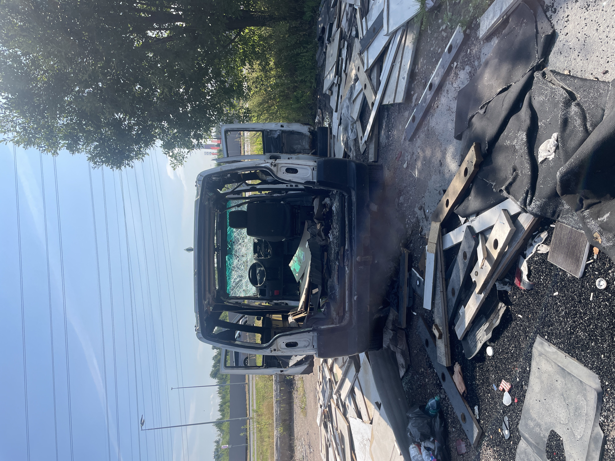 A burnt-out white van is parked on a dirt lot in Pargolovo, Russia. The doors are open, and the interior is gutted, with shattered glass on the windshield and steering wheel. Debris, including wooden planks, plastic, and fabric, is scattered around the van and on the ground. The area is surrounded by vegetation, with trees and bushes visible. Power lines and streetlights are in the background. The sky is clear and blue.