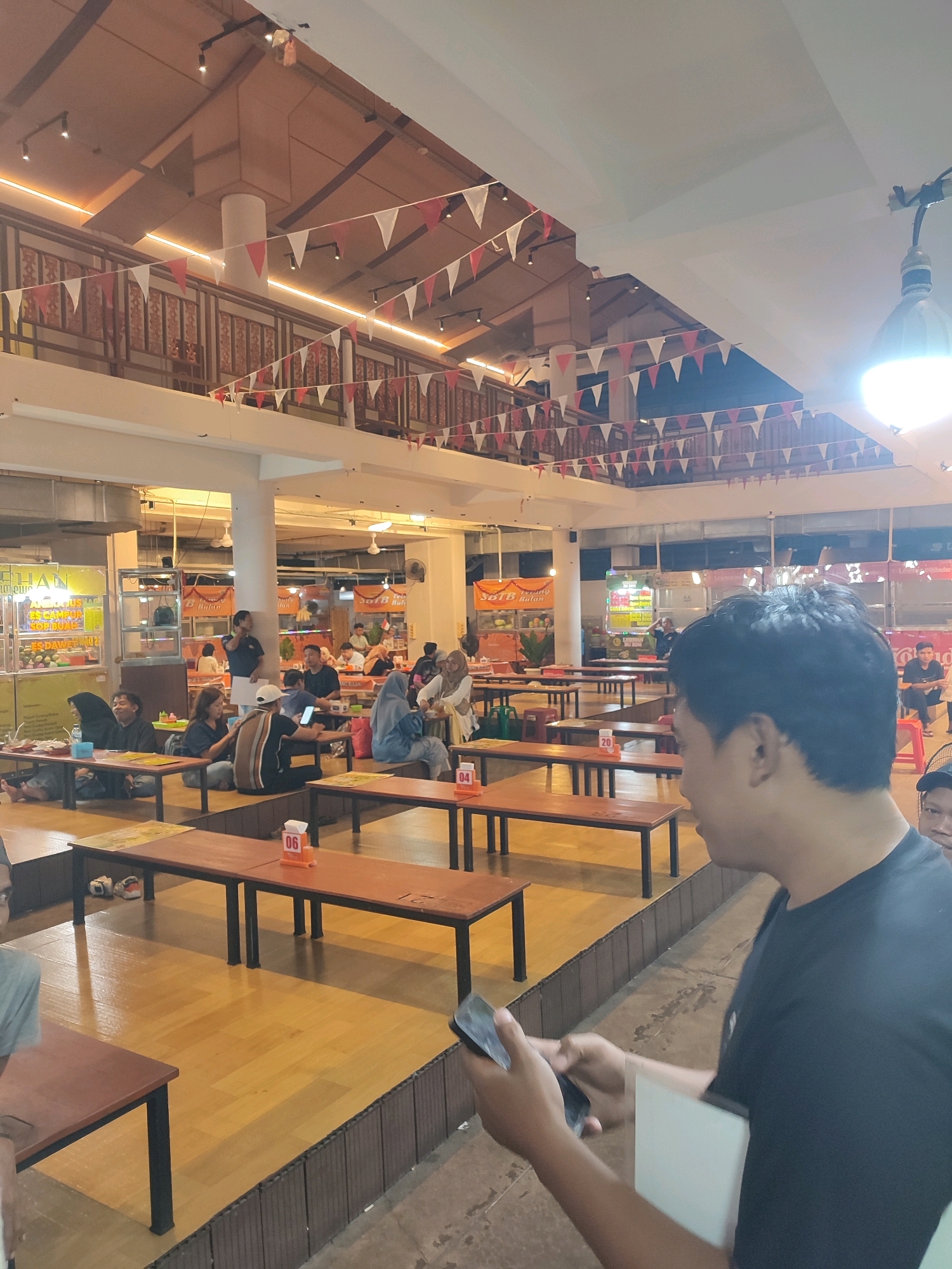 This is an indoor scene at a food court in Yogyakarta, Indonesia, decorated with red and white triangular pennants. The food court has a wooden floor and numerous tables and benches. In the foreground, a man in a black t-shirt is looking at his phone. In the background, people are seated at tables, eating and socializing. There are various food stalls visible, with signage in Indonesian. The lighting suggests it is evening or nighttime.