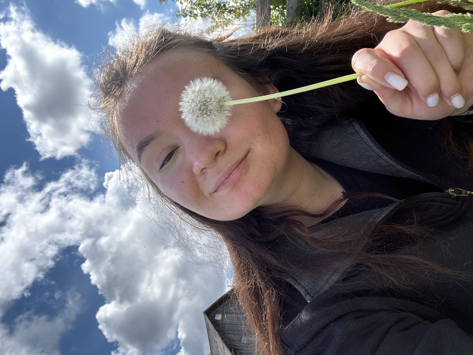 A young woman is holding a dandelion in front of her face. She has brown hair and is wearing a black jacket. The background is a blue sky with white clouds. There is a building in the lower part of the image. The picture was probably taken in Дойная, Russia.