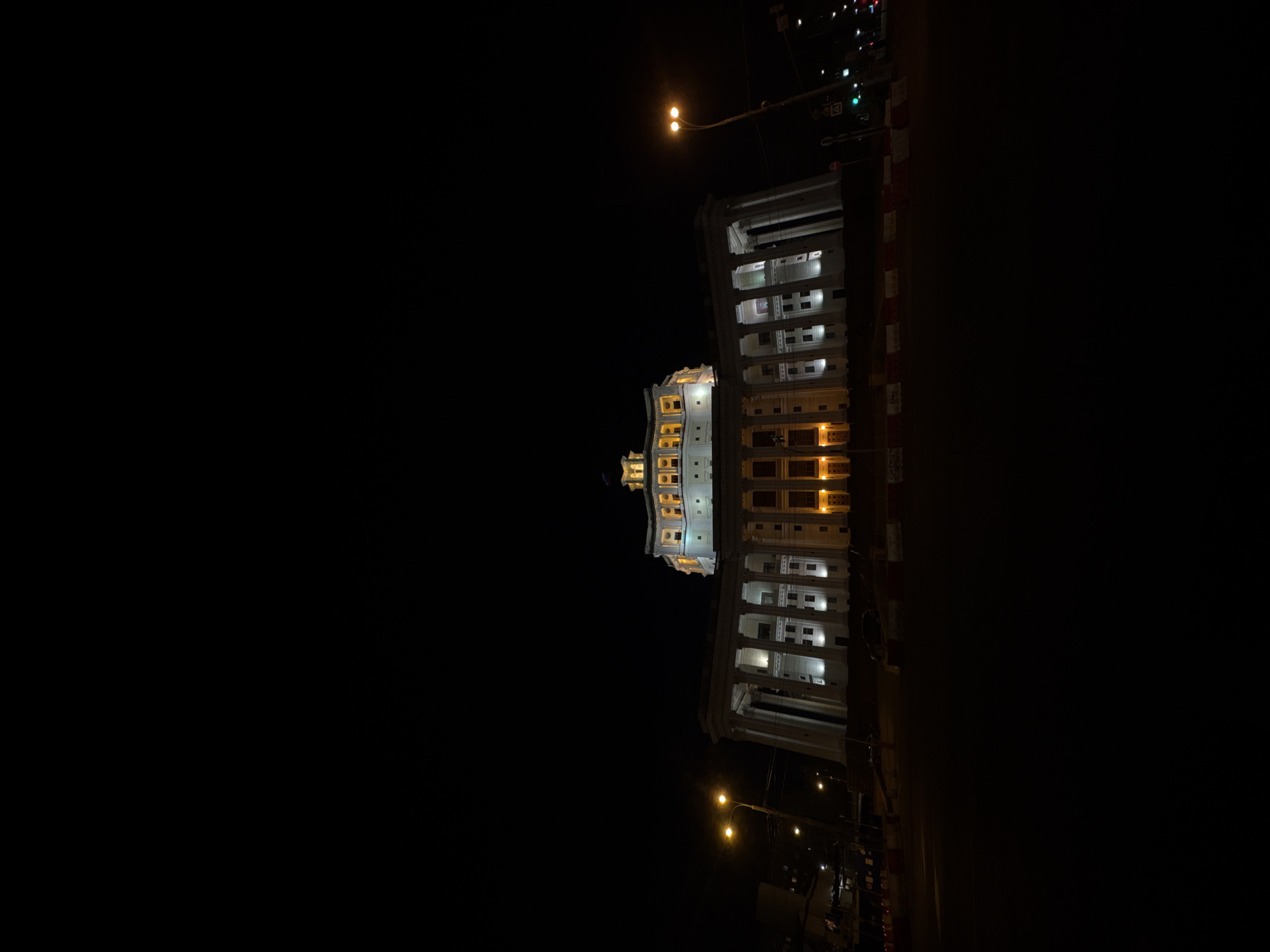 The image shows the illuminated facade of a building at night. The building has a classical architectural style with columns and multiple stories. It is likely located in Moscow, Russia. The environment is dark, suggesting it is late at night. There are streetlights providing additional illumination around the building and the street. There is a street beside the building, divided by barricades.