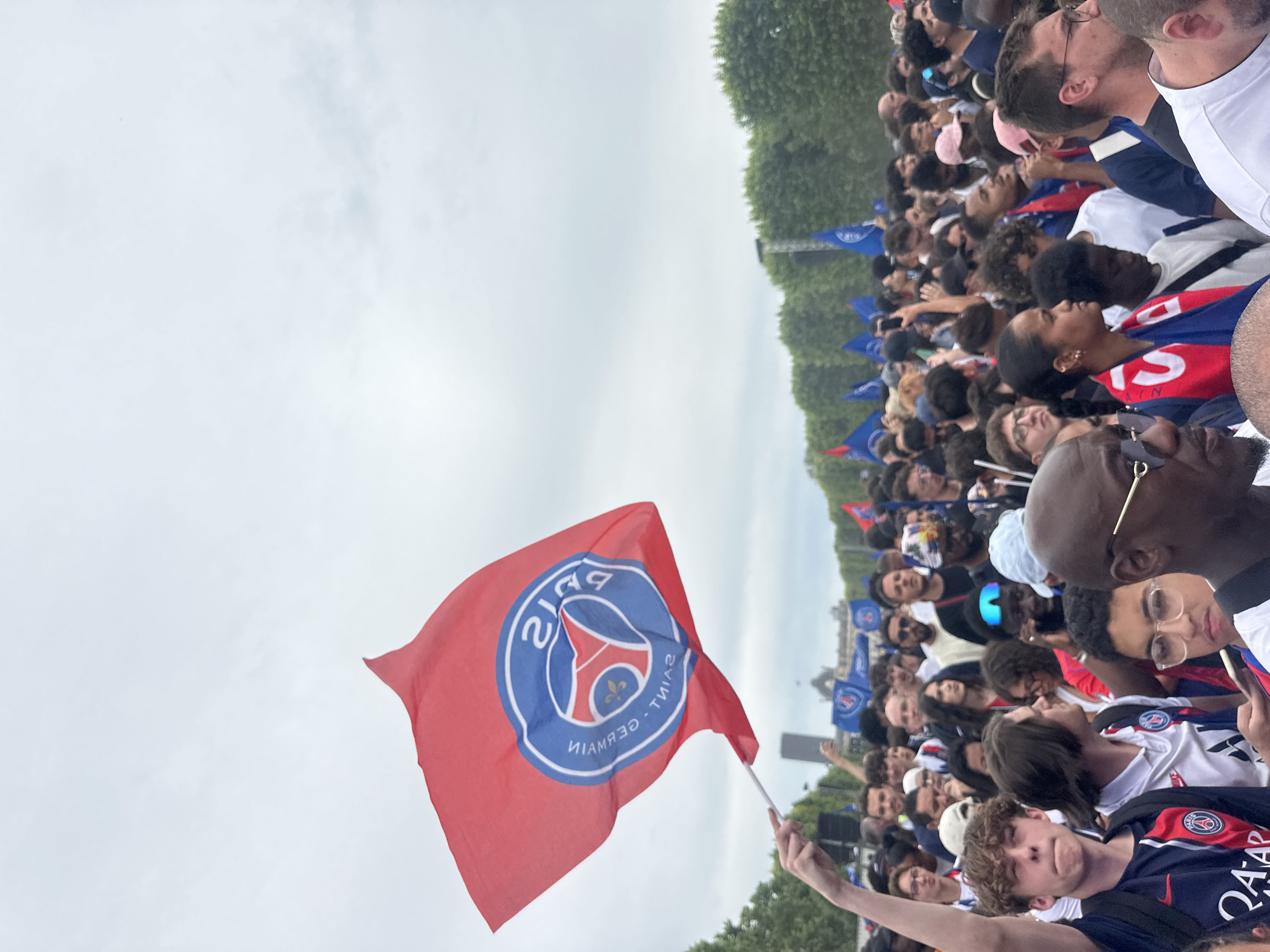 The image shows a large crowd of people outdoors. Many are wearing Paris Saint-Germain (PSG) jerseys and waving PSG flags, suggesting they are fans of the football club. The sky is overcast. Trees are visible in the background.