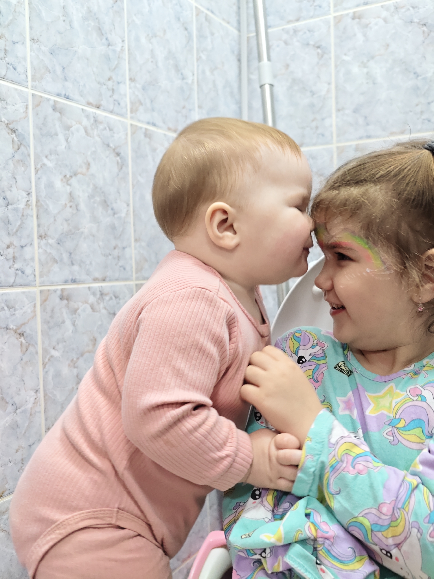 An indoor photograph, likely taken in Chaikovsky, Russia, depicts two young children in close interaction. The background features light blue-grey marbled square tiles with visible grout lines and a vertical silver-colored pipe or fixture in the upper right quadrant. A white object, consistent with a child's potty training seat, is partially visible behind and between the children, with a small pink component visible in the lower right.
The child on the left, an infant or toddler, has light-colored hair and is dressed in a solid pink ribbed long-sleeved bodysuit. This child's face, showing fair skin and visible rosy cheeks, is oriented toward the right, with their forehead making contact with the forehead of the other child.
The child on the right, an older toddler or young child, has light brown hair pulled back with a dark hair tie. This child wears a light blue long-sleeved shirt adorned with colorful patterns of unicorns, stars, and diamonds. A rainbow design is painted over their right eyebrow, and a small pink star-shaped earring is visible in their right ear. The child is smiling, with their gaze directed towards the left child. The left child's left hand is gently placed on the right child's right forearm.