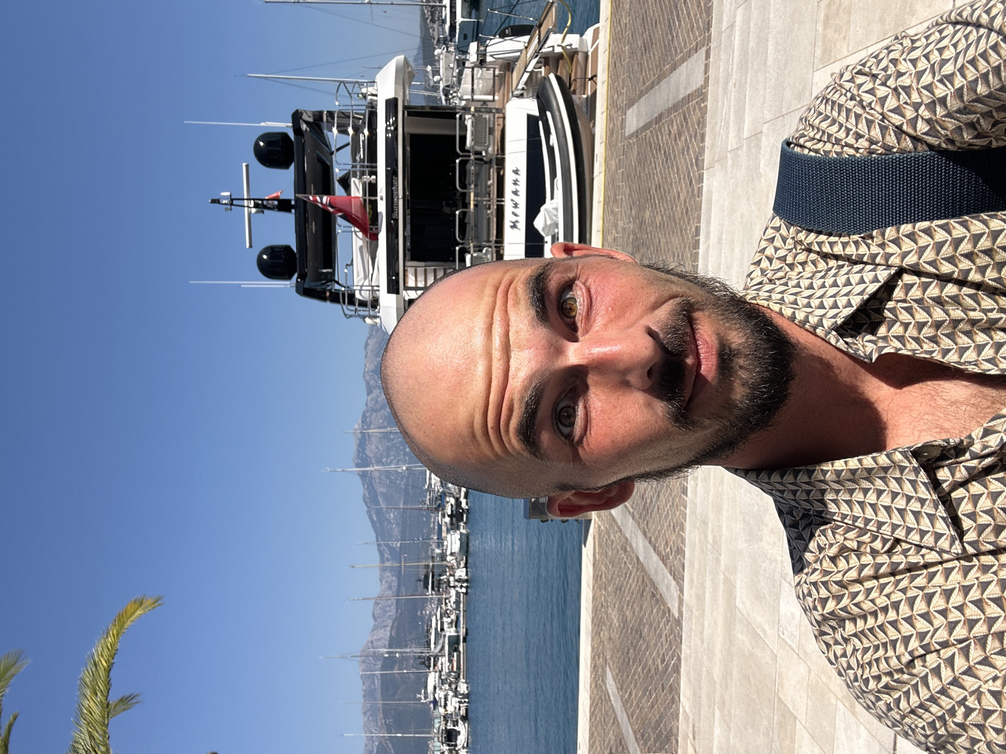 A man with a shaved head and a beard is taking a selfie in a marina. He is wearing a patterned short-sleeved shirt and a dark blue strap across his chest. Behind him, a large white yacht named "MEWHANA" is docked, with a smaller inflatable boat on its stern. Several other sailboats and yachts are moored in the background, with mountains visible beyond them. The sky is clear and blue. The pavement in front of him is made of light-colored stones. The scene appears to be in a sunny, coastal city, likely Herceg Novi, Montenegro.
