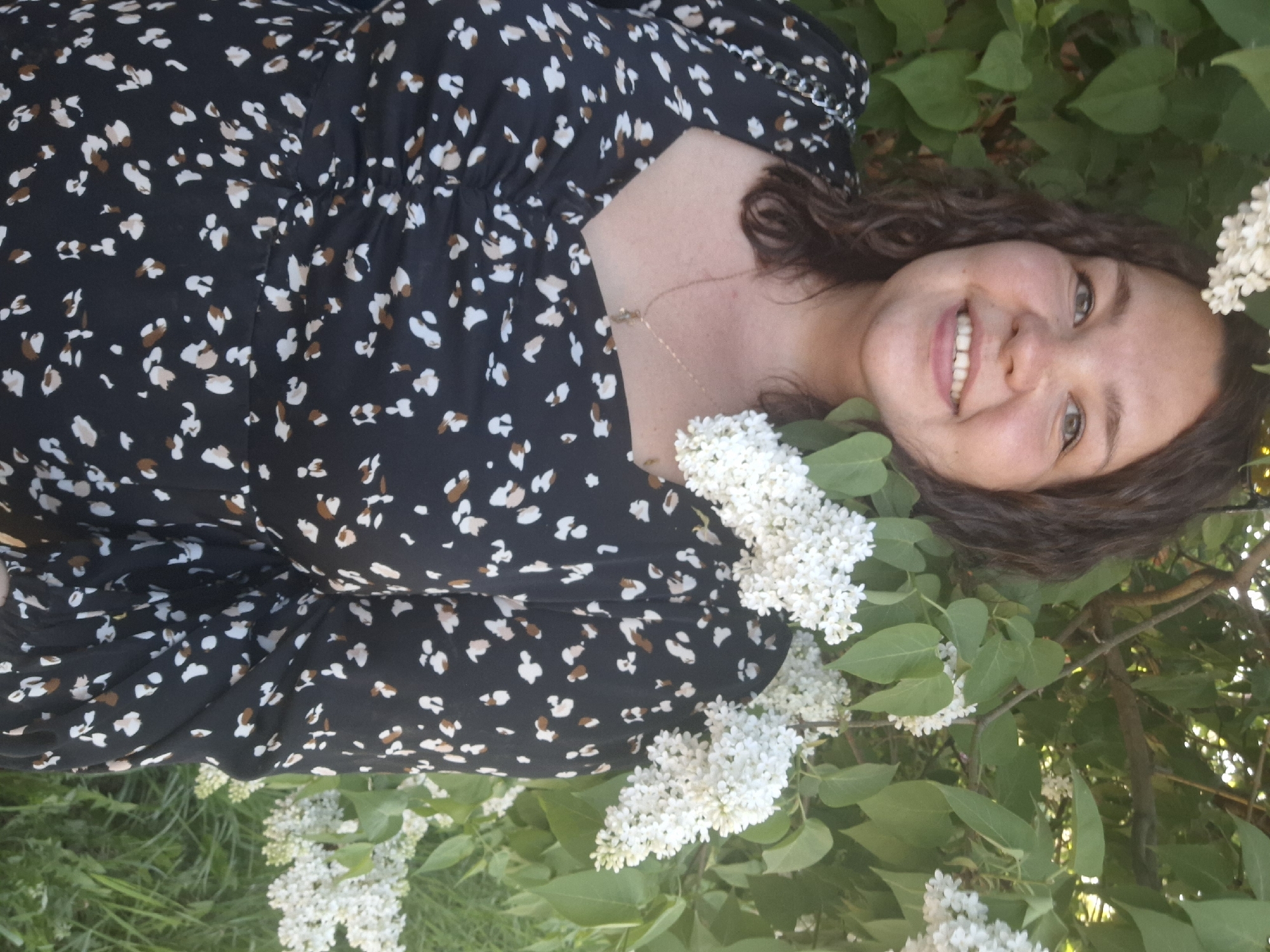 A woman is pictured lying down amongst a blooming lilac bush. She is smiling at the camera. She has brown curly hair and is wearing a black dress with small white and brown speckles. The lilac bush has green leaves and white flowers. It is daytime. The image's location is Chaikovsky, Russia.