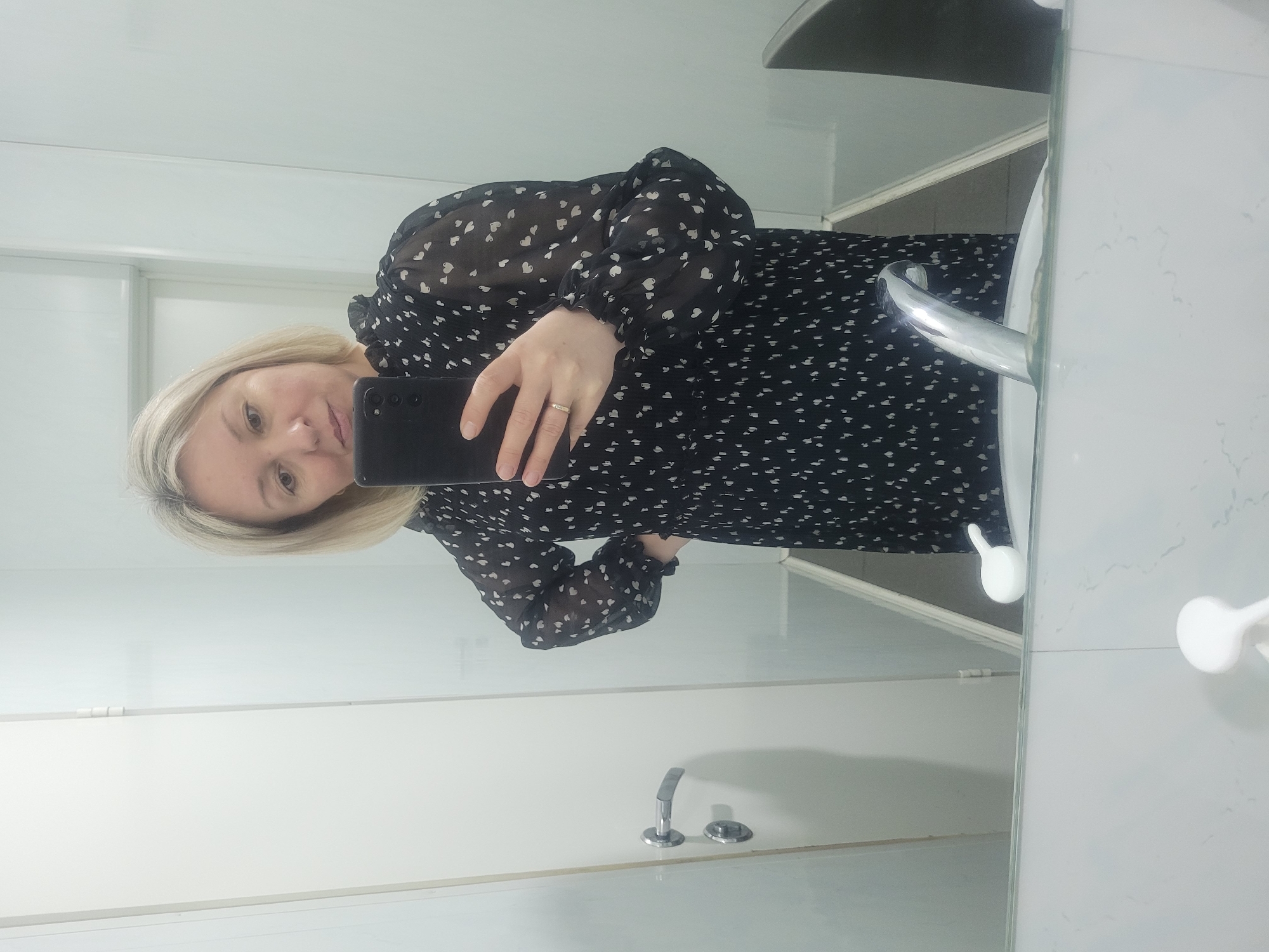 The image shows a person taking a selfie in a bathroom. The person is a woman with blonde hair wearing a black dress with white heart patterns. She is holding a black smartphone, partially obscuring her face. A silver ring is visible on her ring finger. The bathroom features white walls, a sink with a silver faucet, and a soap dispenser. The image is likely taken in a mirror, as indicated by the reversed perspective and the presence of reflections. There is no indication in the image of its location in Izhevsk, Russia.