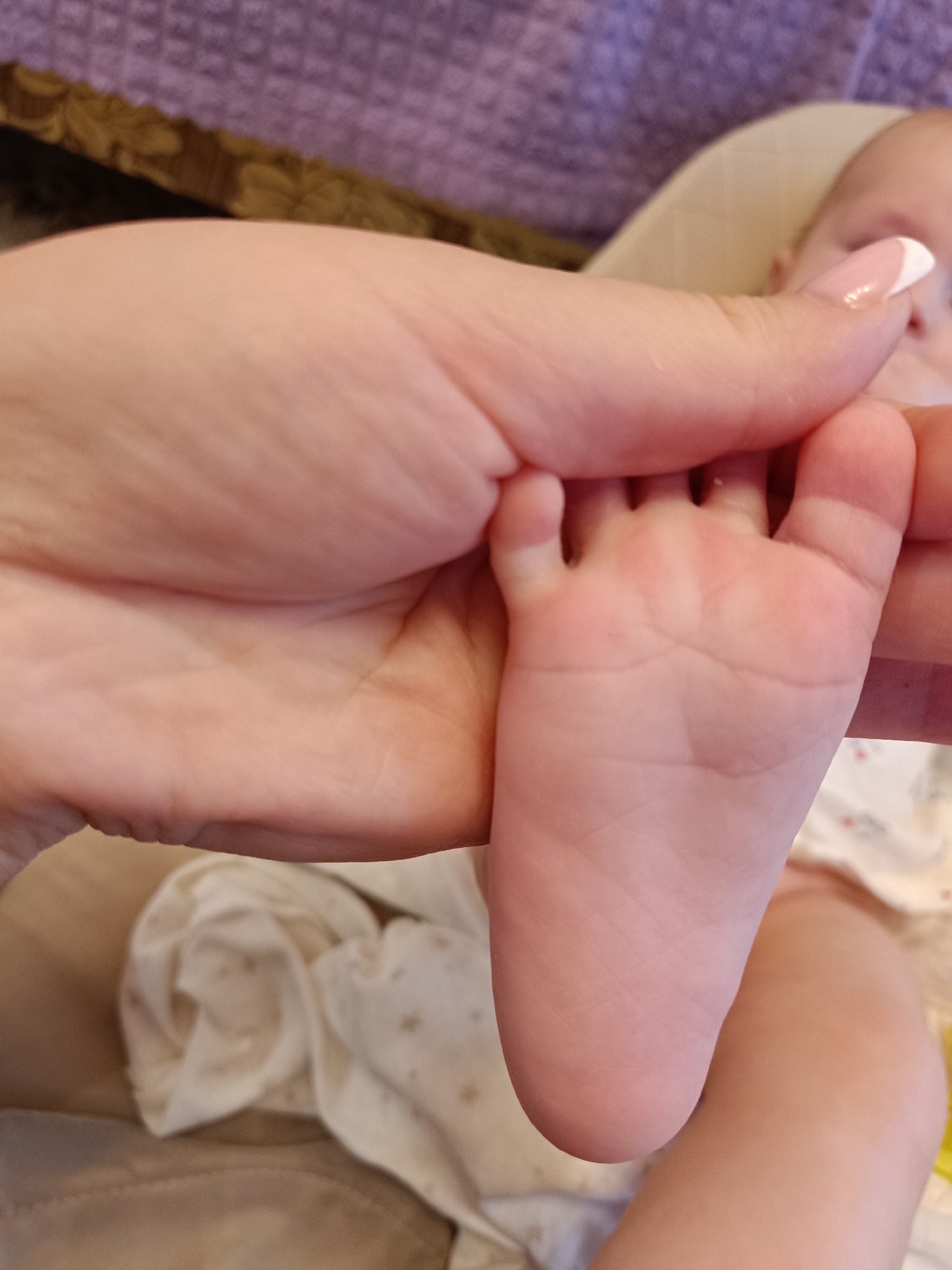 A close-up image shows a baby's foot being held by an adult's hand with a French manicure. The baby is wearing a patterned white outfit. The background includes a purple textured surface and a beige fabric. The location is not discernible from the image.