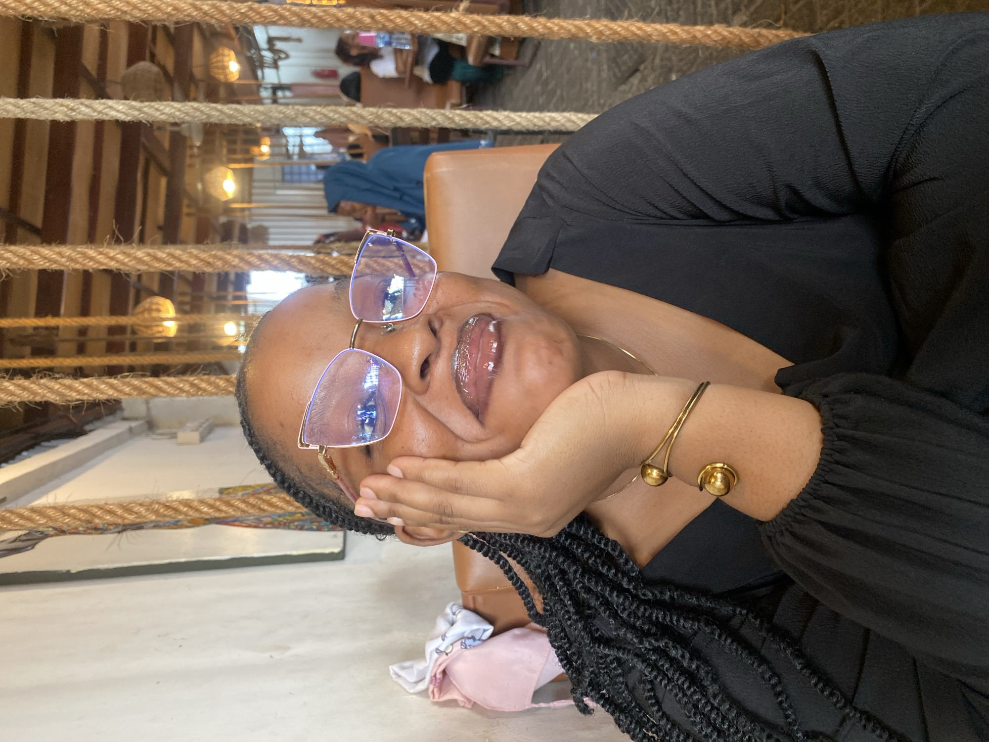 A woman with dark skin is resting her head in her hand while wearing glasses, a black top, and a gold bracelet. She has long, dark braids. The background shows a blurred interior of a cafe, with lights and people indistinctly visible. Ropes hang in the foreground, partially obscuring the background scene. The location appears to be a cafe in Dar es-Salaam, Tanzania.