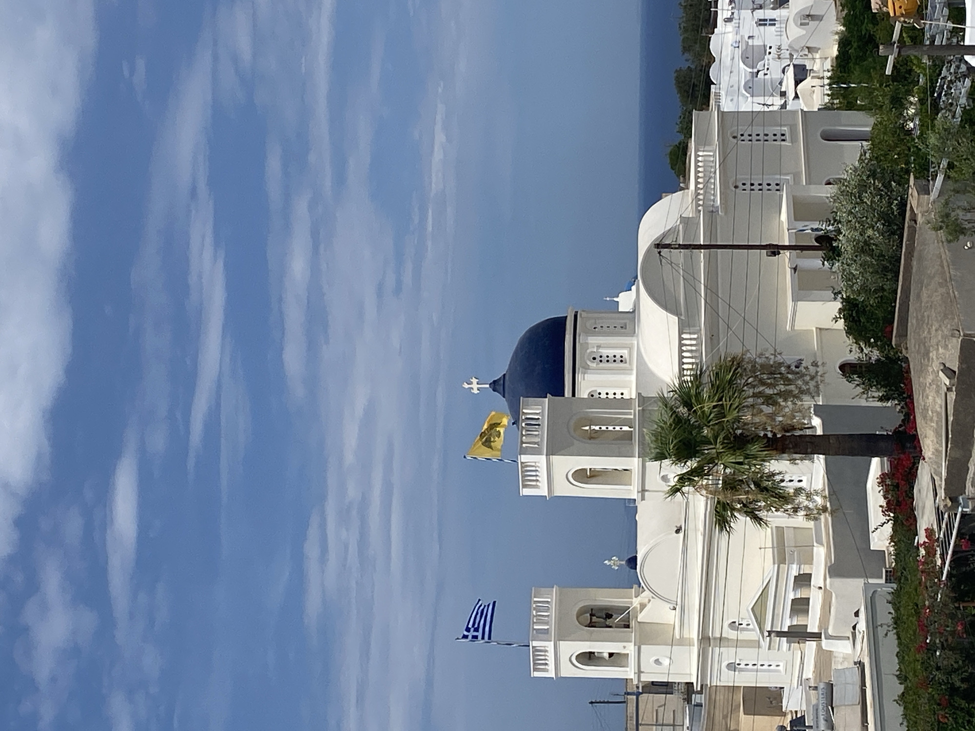 The image depicts a view of the Three Bells of Fira church in Thira, Santorini, Greece, with the Aegean Sea visible in the background under a partly cloudy blue sky. The church features white walls, a prominent dark blue dome, and two bell towers. The Greek flag is flying from the left bell tower, and a yellow flag with a black eagle is displayed on the right tower. A palm tree stands in front of the church, and lush green vegetation and red flowers are visible in the foreground. The surrounding architecture is characteristic of Santorini, with white buildings and domed roofs. The scene is bathed in daylight.