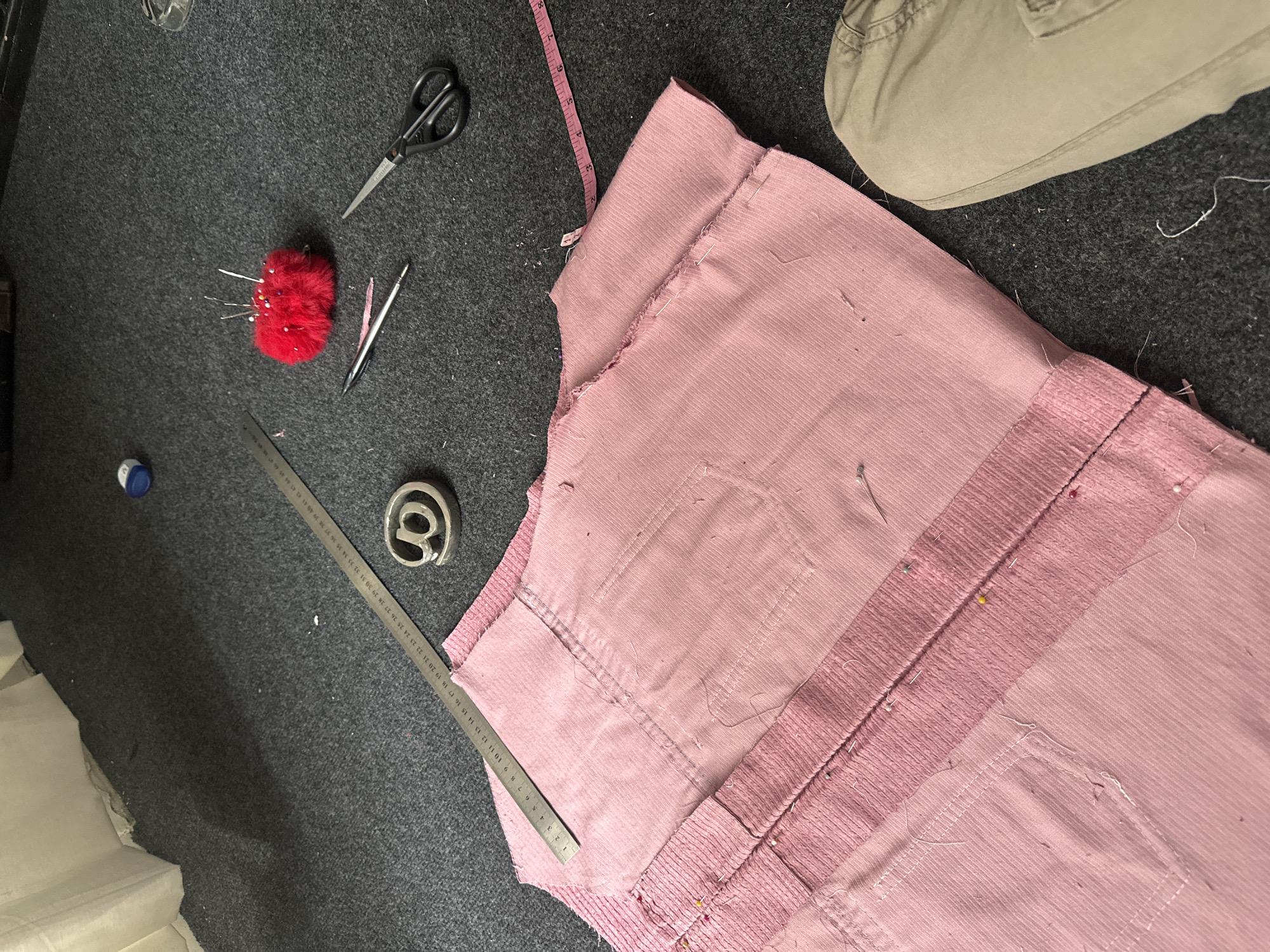 The image depicts a sewing project in progress on a dark gray, textured surface, likely a carpet or fabric. The main subject is a partially assembled garment made from pink corduroy material. It shows various elements associated with sewing, including:
* A red pincushion filled with sewing pins
* A pair of scissors
* A seam ripper or similar tool
* A metal ruler
* A measuring tape
* A symbol (@)
The pink garment is pinned together in places, suggesting the pieces are being prepared for sewing. The presence of the ruler, measuring tape, and scissors further reinforces the notion that the image captures a sewing project at some stage of development.
There is no indication of the location being in Buenos Aires, Argentina, or any evidence of people present in the image. The scene is focused solely on the materials and tools involved in the sewing project.