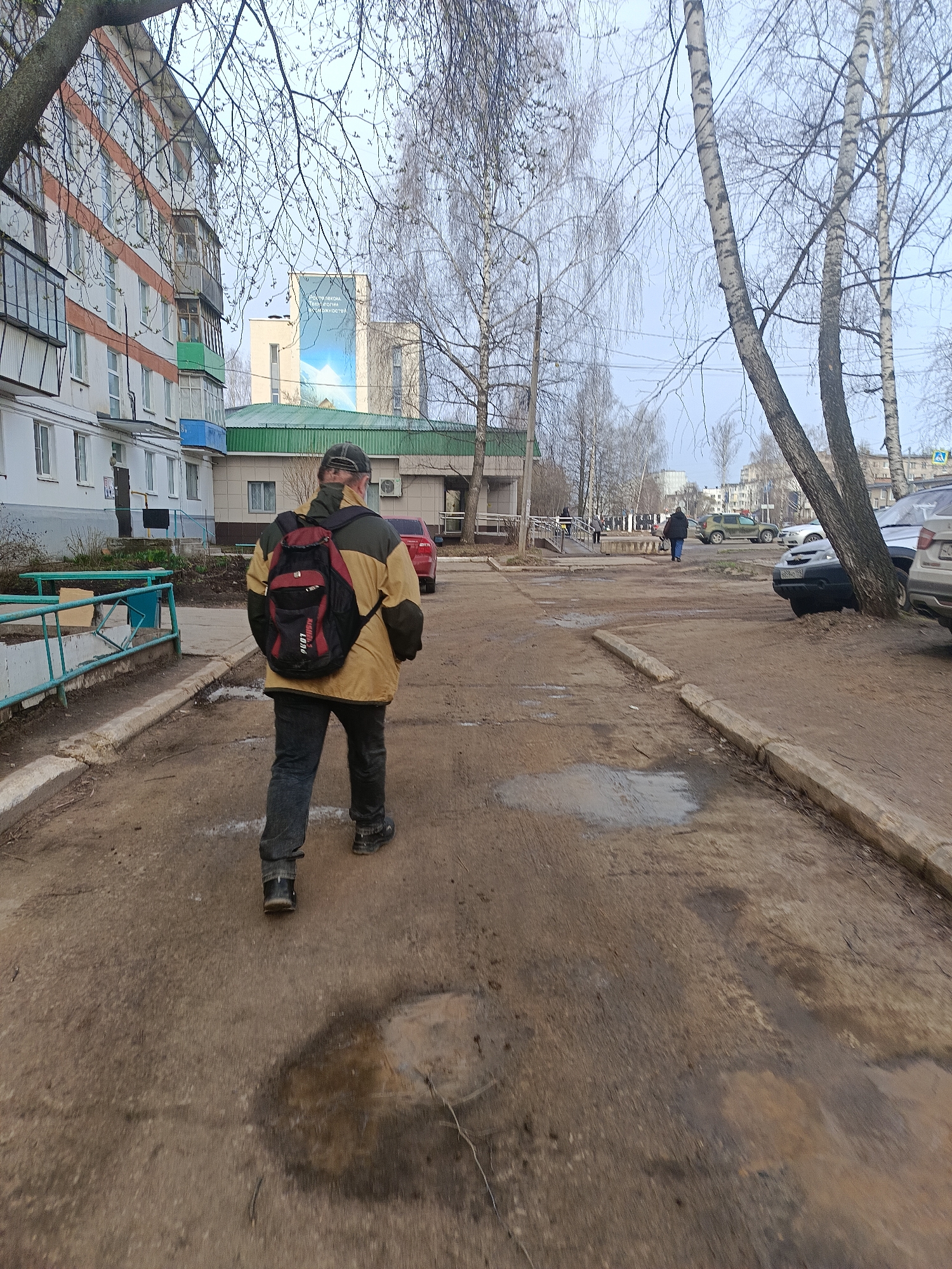 A man wearing a khaki jacket, black pants, a baseball cap, and a red and black backpack is walking away from the viewer down a dirty, cracked sidewalk in the city of Chaikovsky, Russia. To the left is a multi-story apartment building with white walls and orange trim. Adjacent to the building is a shorter green-roofed structure. A red car is parked near the green building. Several bare trees are visible. In the distance, other people are walking and multiple cars are parked. The sky is overcast. Puddles are on the ground suggesting recent rain.