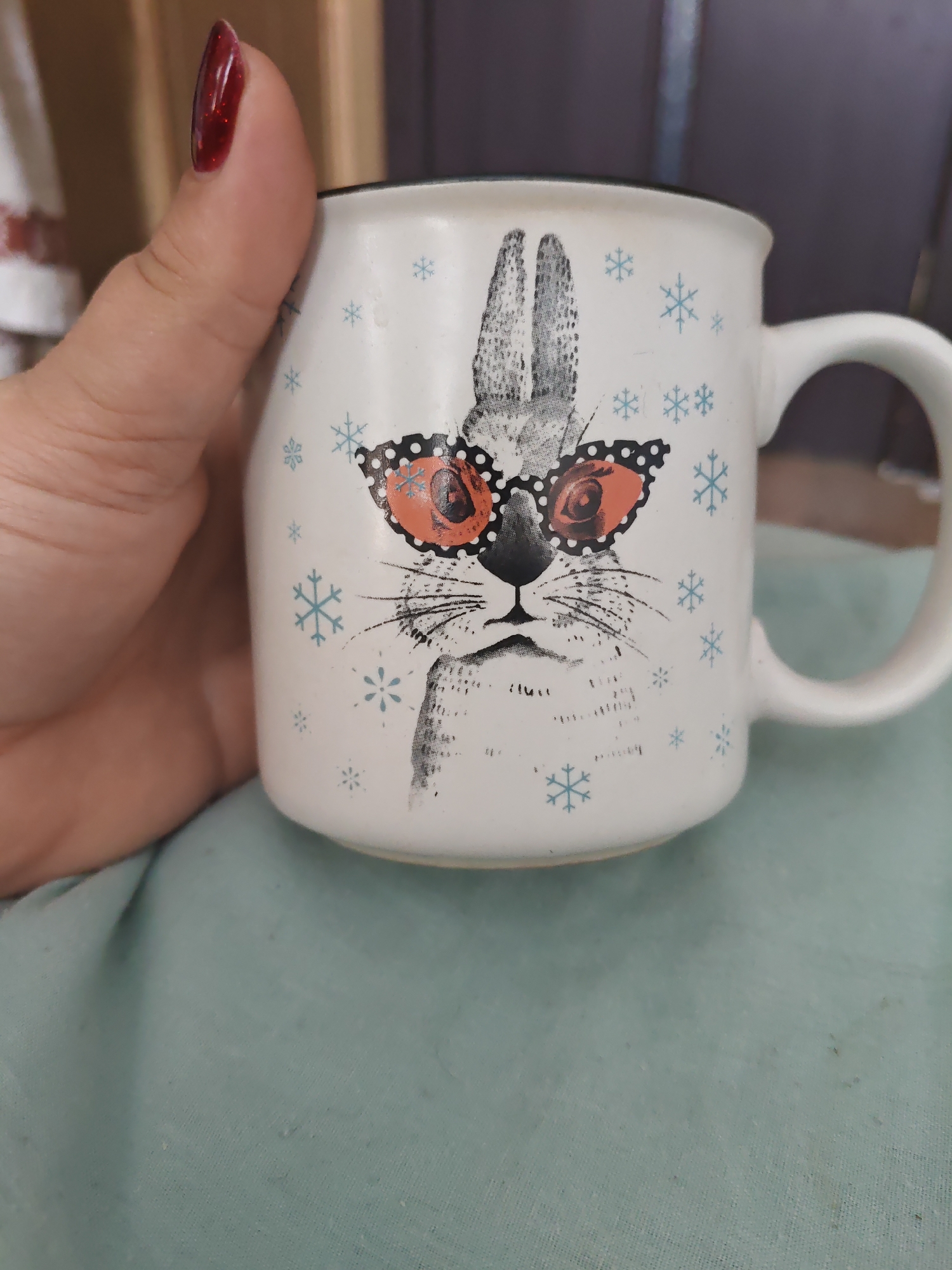 A close-up, eye-level shot shows a hand holding a white ceramic mug with a black rim. The mug features a printed design of a rabbit wearing oversized, orange-tinted sunglasses with black polka dots. Small blue snowflakes are scattered around the rabbit illustration. The background is blurred, with hints of wooden shelving and a dark surface. The scene is set against a pale teal fabric. The image is dated to 2023-11-03. No specific location in Unarokovo, Russia, is visible or discernible from the image.