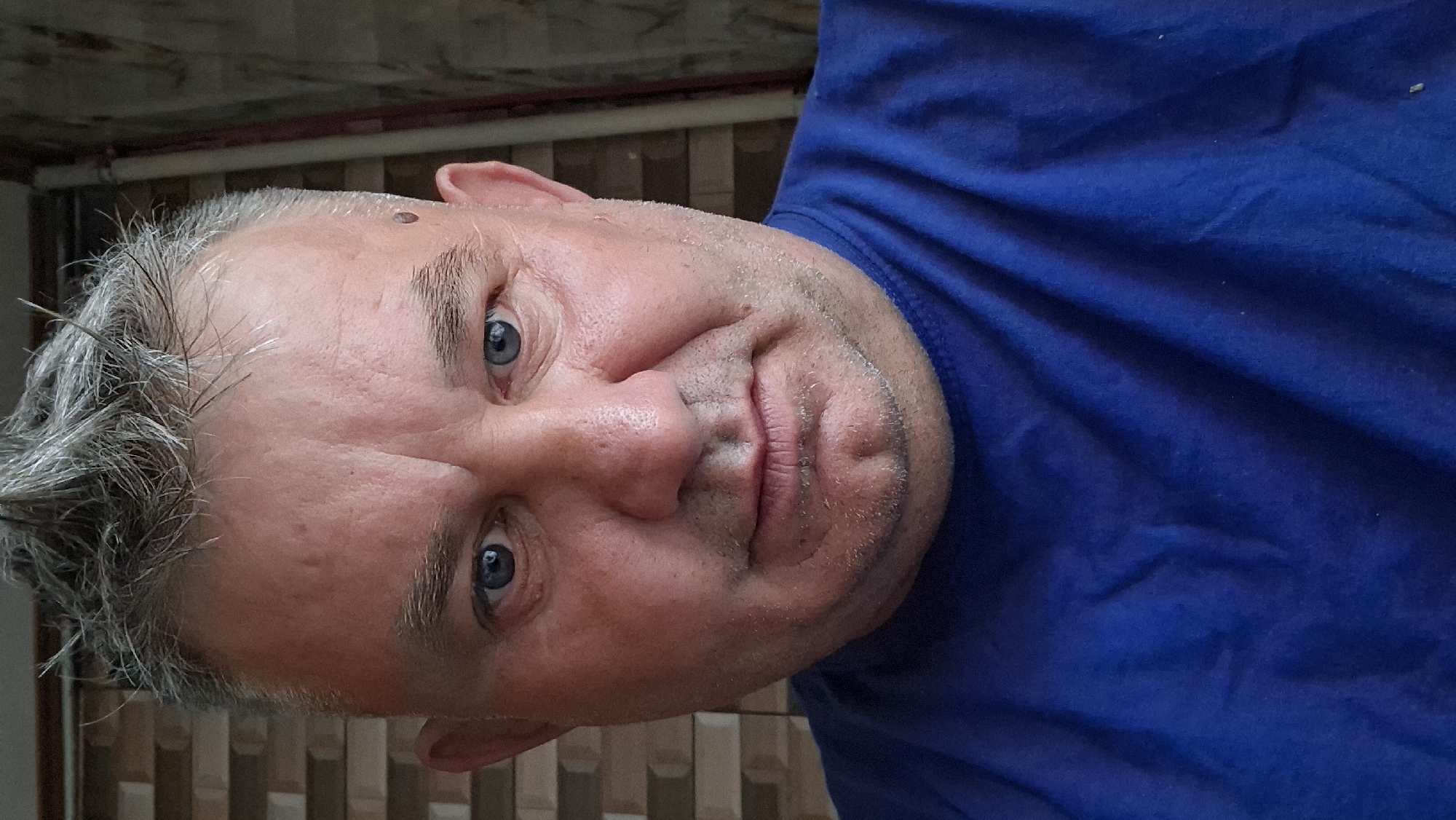 A man with short, spiky, grey hair and blue eyes is looking directly at the camera. He has fair skin with some redness, visible pores, and a mole on his right temple. He is wearing a blue t-shirt. The background consists of a brick wall and a vertical pipe. The image appears to be a selfie taken indoors. The setting is likely a residential area in Voinova, Moldova.