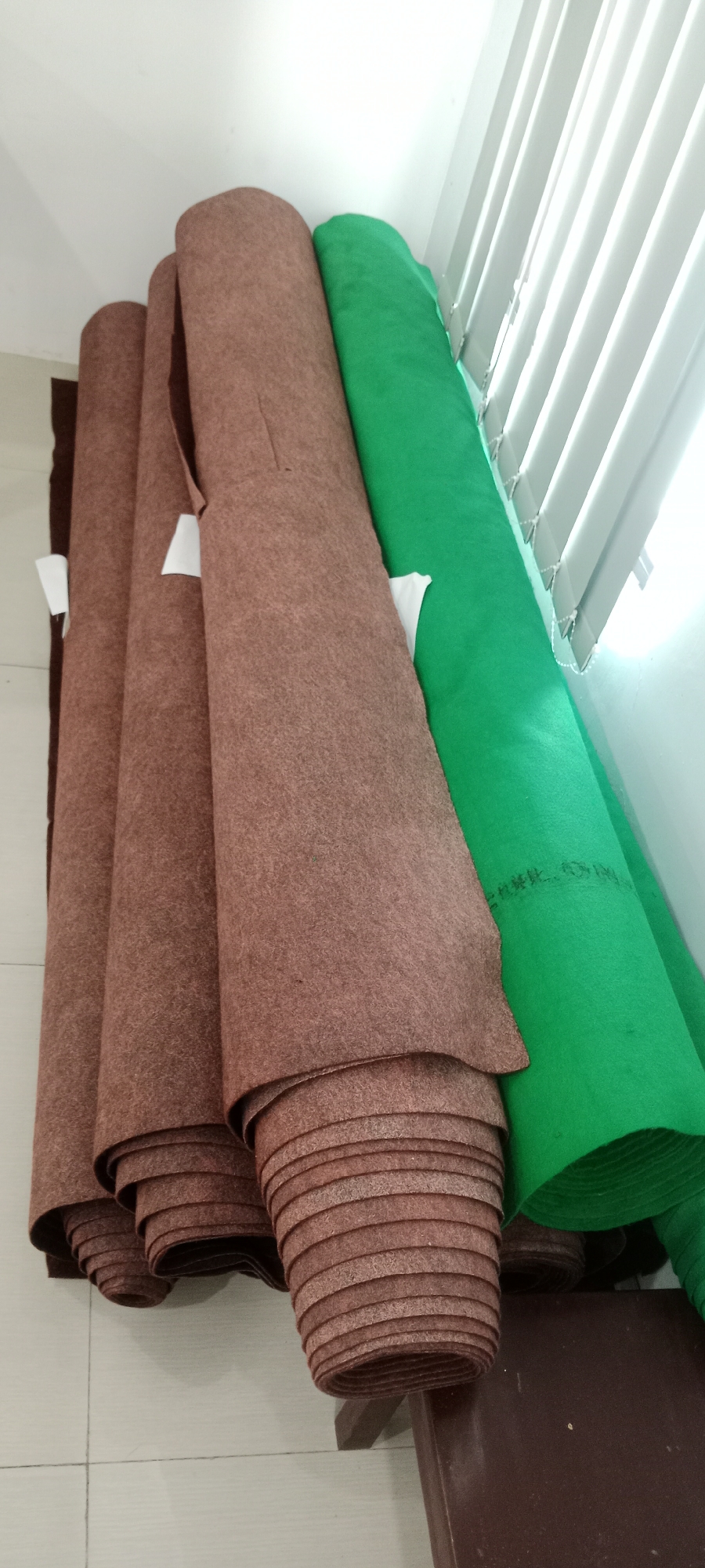The image shows several rolls of felt fabric, one of which is bright green and the others are a reddish-brown color. The rolls are leaning against a wall with white vertical blinds. The flooring appears to be tiled. A dark brown surface, possibly a bench or shelf, is visible at the bottom right of the image supporting some of the rolls. The lighting suggests an indoor environment, possibly a storage area or retail space. There are no people or animals present in the image. The scene is static and depicts stacked rolls of fabric. The location is specified as Tuban, Indonesia, but this information cannot be visually confirmed from the image itself.