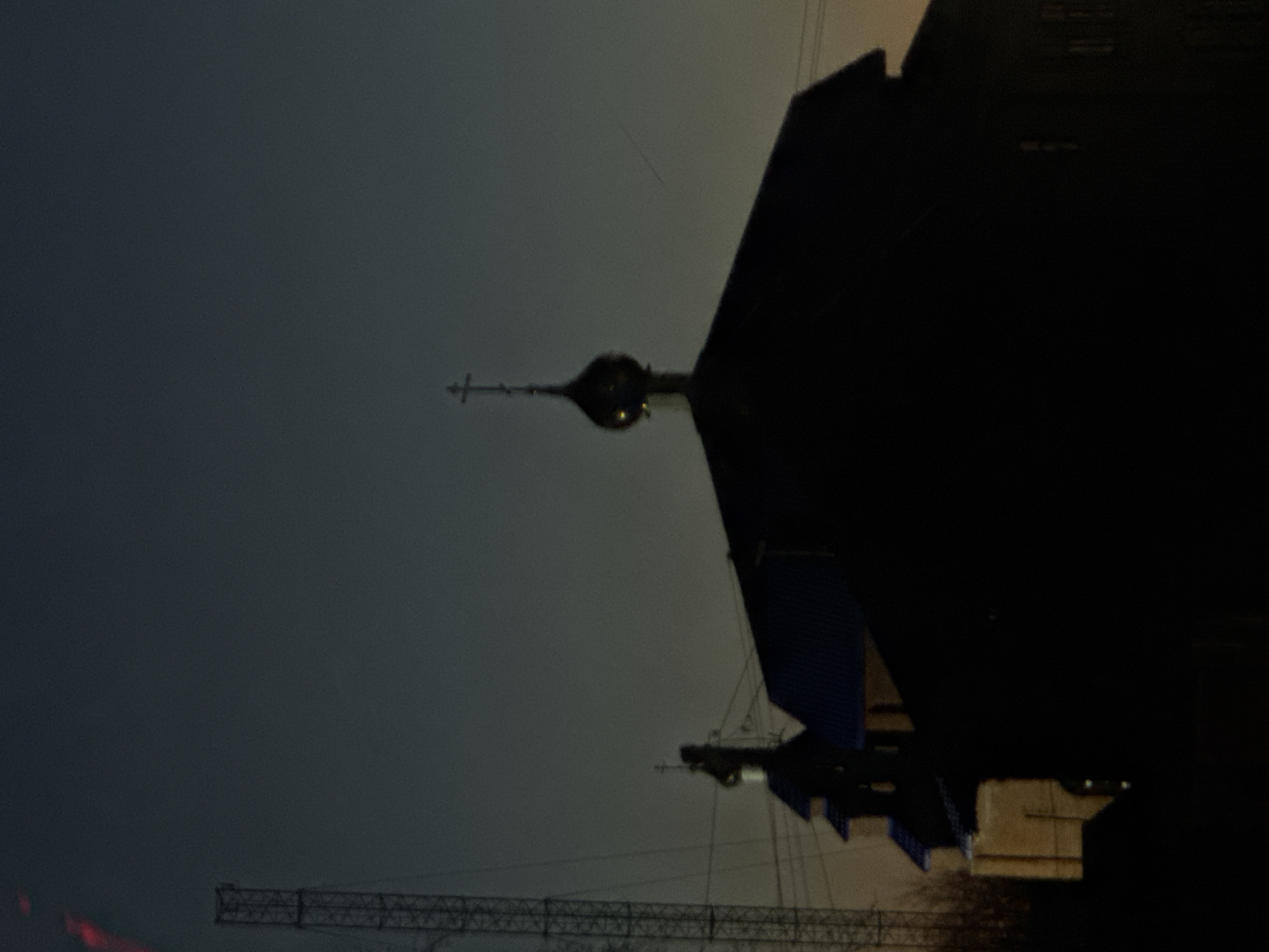 The image, rotated 90 degrees counterclockwise, depicts a dark urban scene in Фоки, Russia, dominated by a silhouette of a building with a prominent Orthodox-style dome and cross. A large construction crane is visible in the lower portion of the image, suggesting ongoing construction or development in the area. The sky is a uniform dark grey, likely indicating twilight or nighttime. Additional structures are partially visible behind the main building.