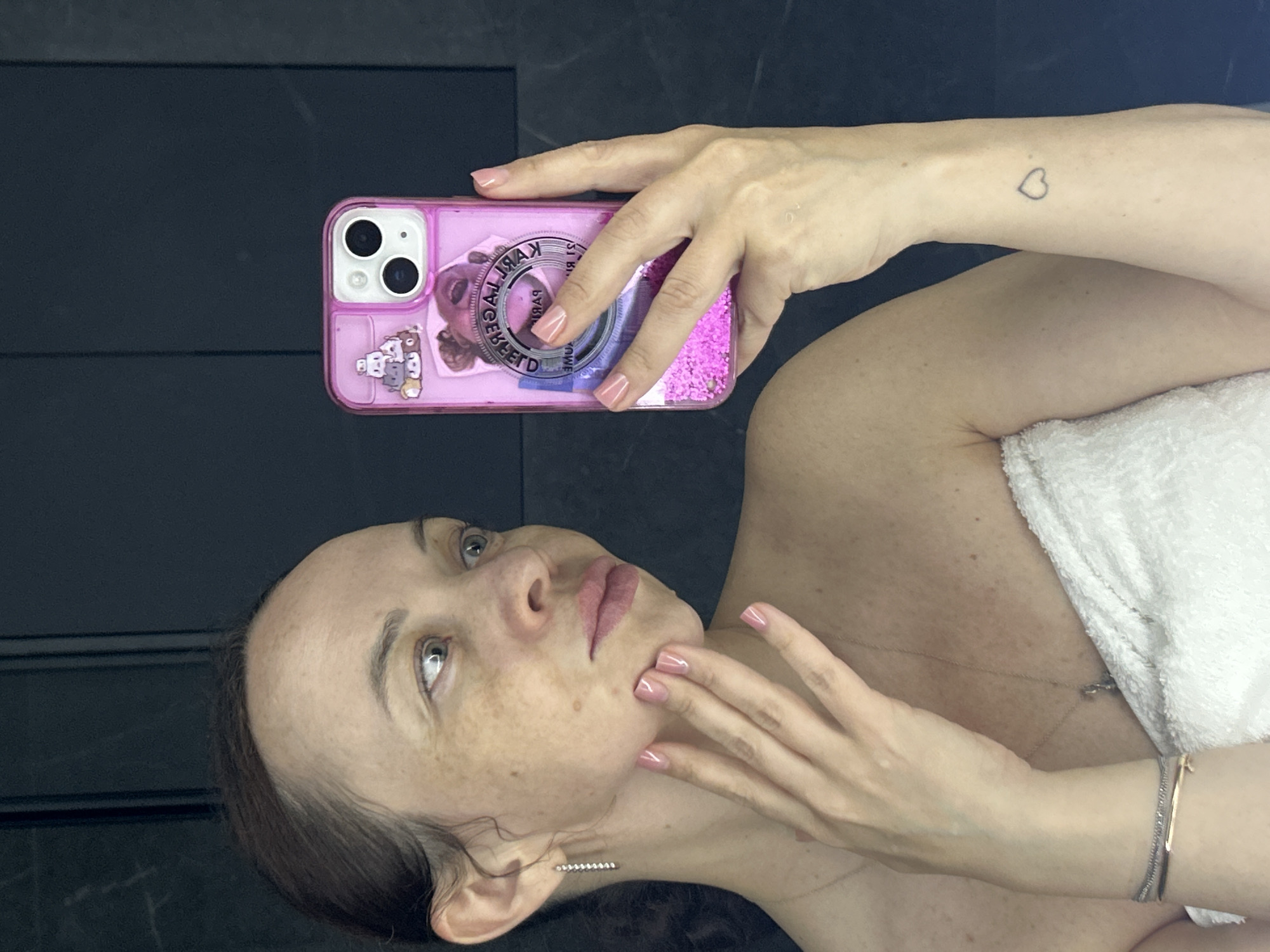 This image depicts a female individual taking a selfie in Yekaterinburg, Russia. The subject, an adult female with brown hair pulled back, is seen from the chest up, wrapped in a white towel. Her skin displays numerous freckles on her face, shoulders, and arms. She wears a silver dangle earring in her visible left ear, a thin gold necklace with a small pendant, and multiple bracelets (one gold-colored, one silver-colored with stones) on her left wrist. A small outline tattoo of a heart is visible on her left forearm. Her fingernails are painted with a light pink, glossy polish.
She is holding a pink smartphone, likely an iPhone given the dual camera configuration on its back, in her right hand. The phone case is transparent pink with floating pink glitter and features stickers, including a stack of cartoon animals and a mirrored circular logo appearing to read "KARL LAGERFELD PARIS." Her left hand is placed with fingers gently touching her left cheek/jawline. She is looking to her right (viewer's left) with a slight side glance and pursed lips. The background is a dark, solid surface, possibly a wall or paneling, with subtle vertical elements, indicating an indoor setting.