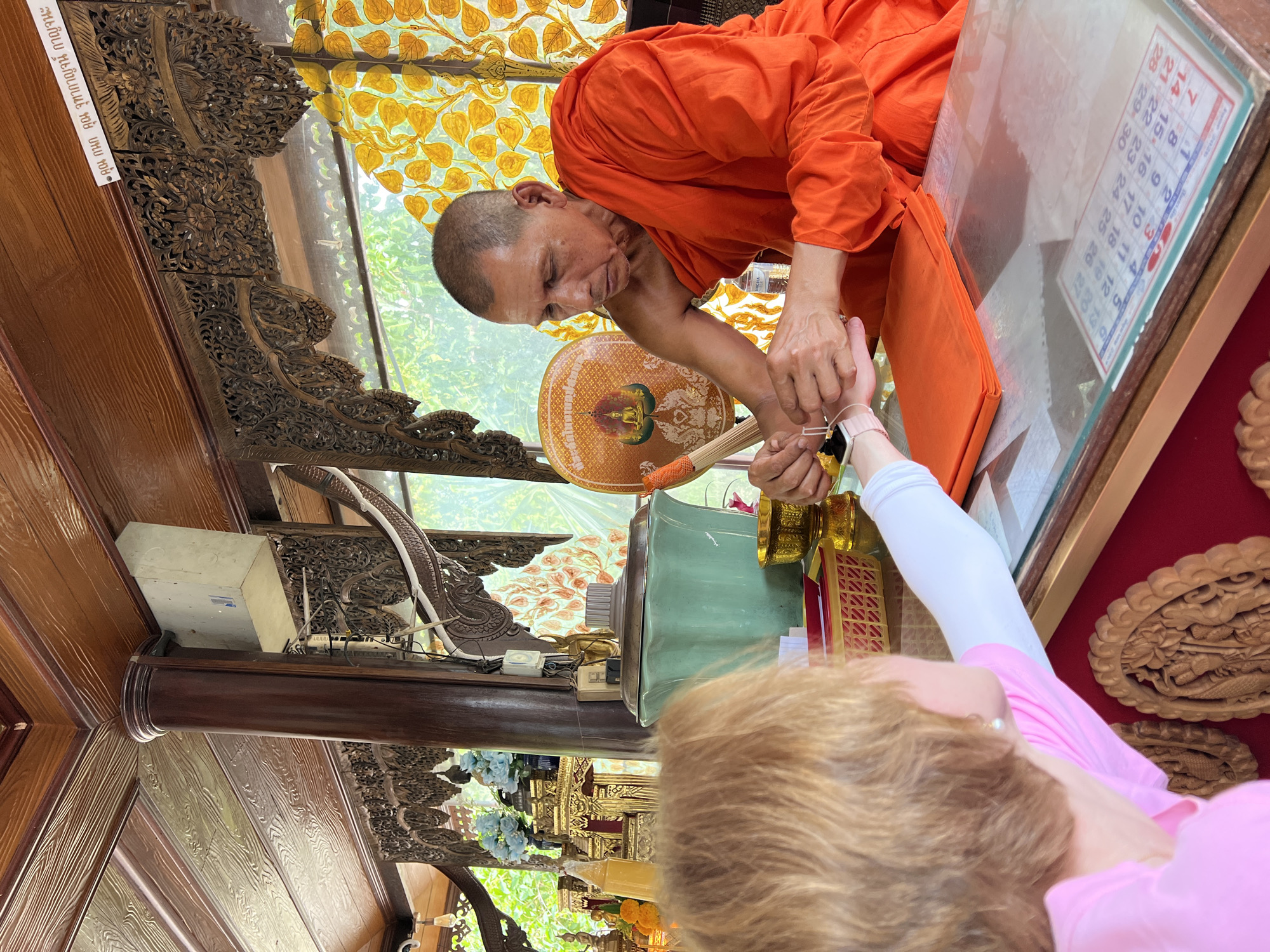 In Ban Bang Rak, Thailand, a Buddhist monk in orange robes is performing a ritual on a woman's wrist. The monk is holding a thin string and a black object, possibly an amulet, while the woman extends her arm. They are situated in a wooden structure with ornate carvings and a yellow, patterned window or screen. A table between them displays a folded orange cloth and a calendar. In the background, there are religious artifacts, including golden statues and blue flowers, and an open window showing greenery. The scene appears to be within a temple or religious site.