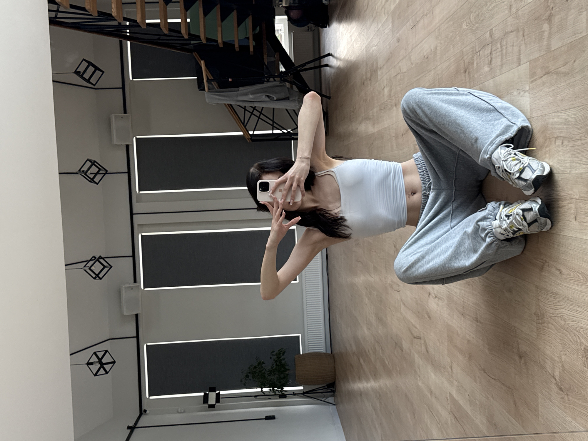 A young woman is taking a selfie in a room with wooden floors. She is wearing a white tank top and grey sweatpants. Her legs are crossed and she is holding a white iPhone up to take the picture. She has black hair and is wearing white sneakers with yellow laces. In the background, there are windows with blinds, a plant in a basket, and a staircase. The scene appears to be indoors during the daytime.