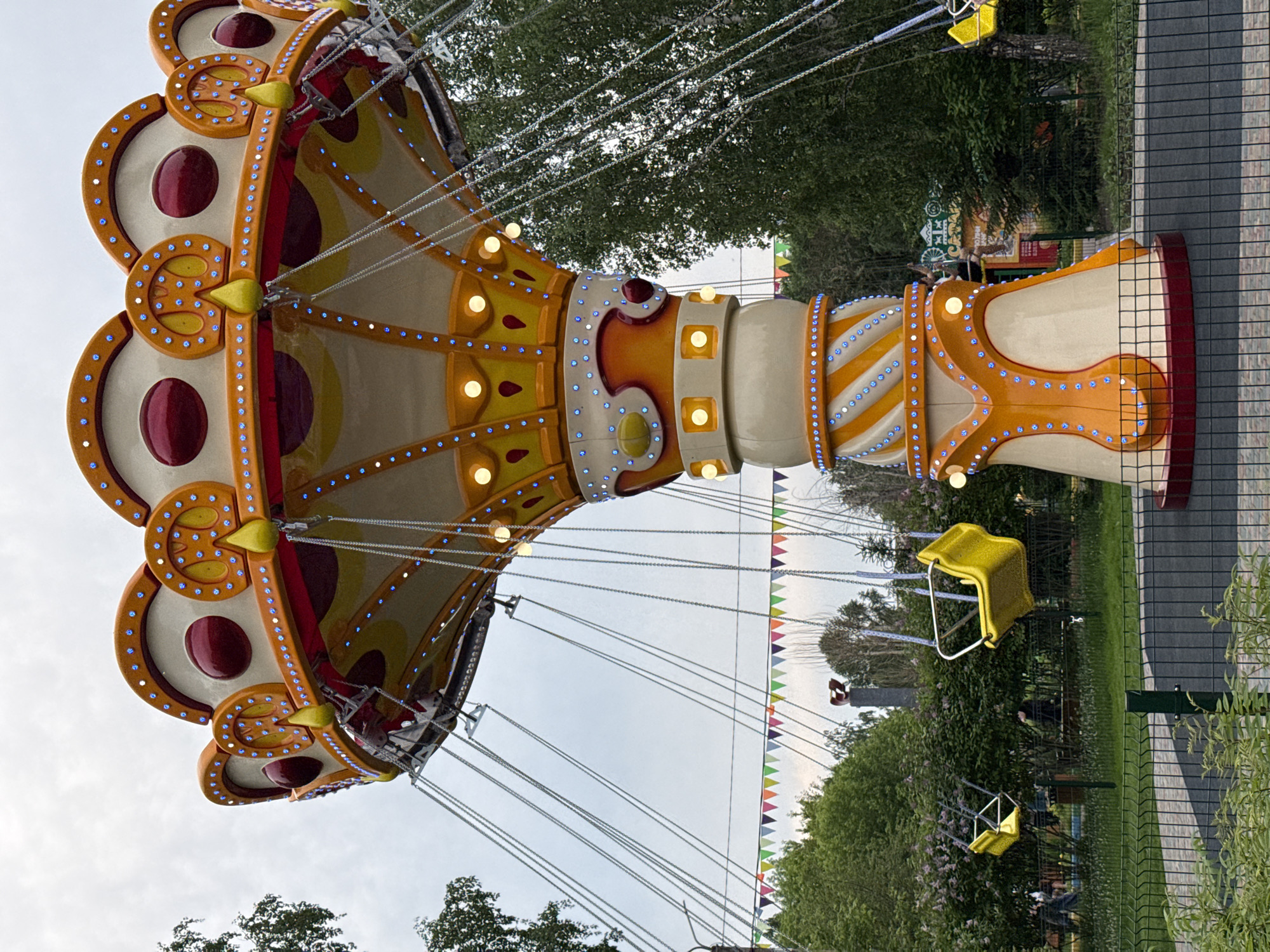 This is a medium shot, eye-level photograph of a swing ride carousel at an amusement park. The ride is predominantly orange and yellow, with decorative blue lights and red accents. Several yellow swings hang from silver chains, some occupied by unseen individuals. The ride is situated in a green, tree-filled park with a grey, cloudy sky overhead. In the background, a series of colorful triangular flags are strung between trees. The area is enclosed by a dark green fence. The location is identified as the "Park of the City of Krasnoyarsk, Russia." The overall atmosphere is one of outdoor recreation.