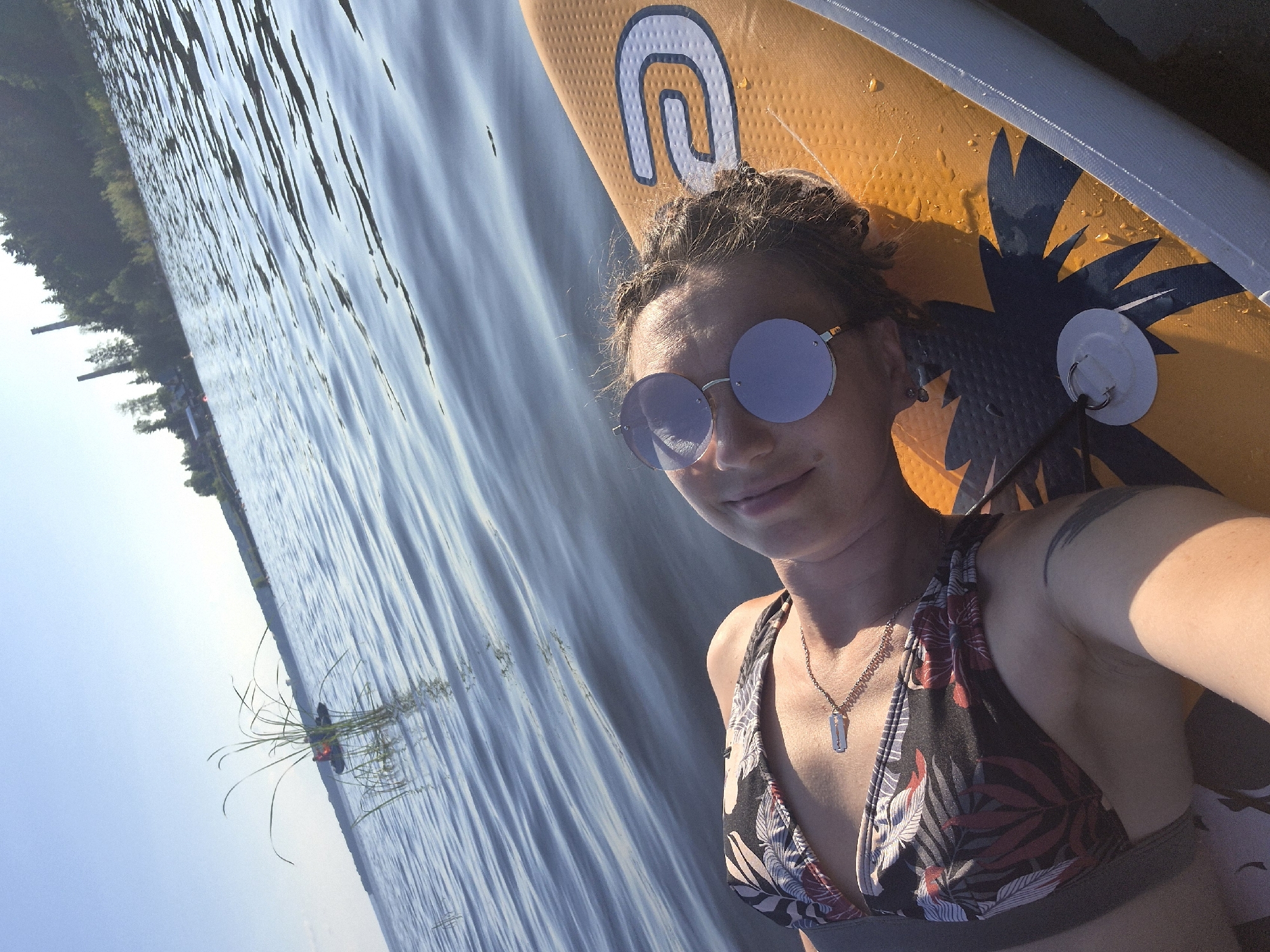 A woman is lying on an orange paddleboard in a body of water, taking a selfie. She is wearing a patterned bikini top and round, light purple sunglasses. Her hair is styled in braids and pulled up. She is smiling slightly, and her expression suggests a relaxed or happy mood. A silver necklace with a rectangular pendant is visible around her neck. There is a tattoo on her right shoulder. The paddleboard has a large "U" logo and a dark blue palm tree design. Water droplets are visible on the surface of the paddleboard, indicating it has been in the water. The background shows rippling water and a distant shoreline with trees. The sky is clear and blue. The lighting suggests it is daytime, likely late afternoon or early evening due to the warm sunlight. The scene appears to be a recreational activity on a lake or calm river.