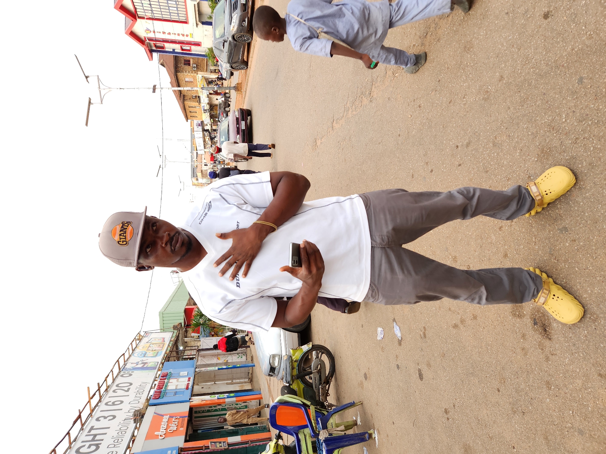 In an outdoor, urban street scene, a man wearing a white t-shirt with "GIANTS" branding on a baseball cap and grey pants stands in the foreground, holding a mobile phone in his right hand and resting his left hand on his chest. He is wearing yellow clogs. To his right, a young boy in a light blue outfit walks away from the camera. In the background, various buildings, shops, and vehicles line the street. A motorcycle with two plastic chairs is parked to the left of the frame. Several other individuals are visible walking in the street. The ground is paved asphalt.
