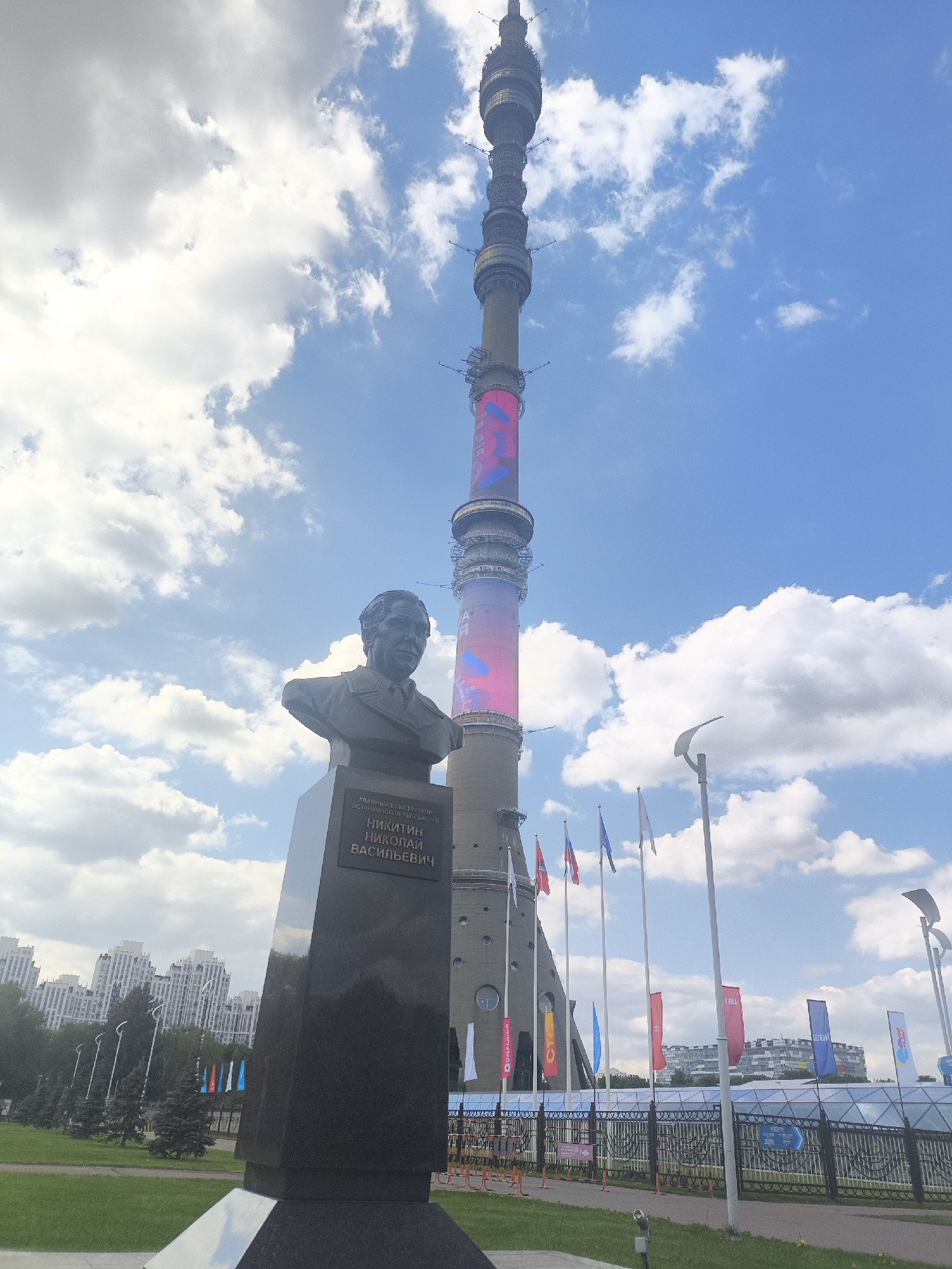 The image shows a bust of Nikolai Nikitin, a Russian structural engineer, located in front of the Ostankino Tower in Moscow, Russia. The bust is mounted on a tall, dark-colored pedestal with text in Russian. The Ostankino Tower, a tall television and radio tower, rises in the background, partly obscured by clouds. Several flags are flying in the middle ground behind the bust. In the far background, residential buildings are visible along the horizon under a sky with scattered clouds.