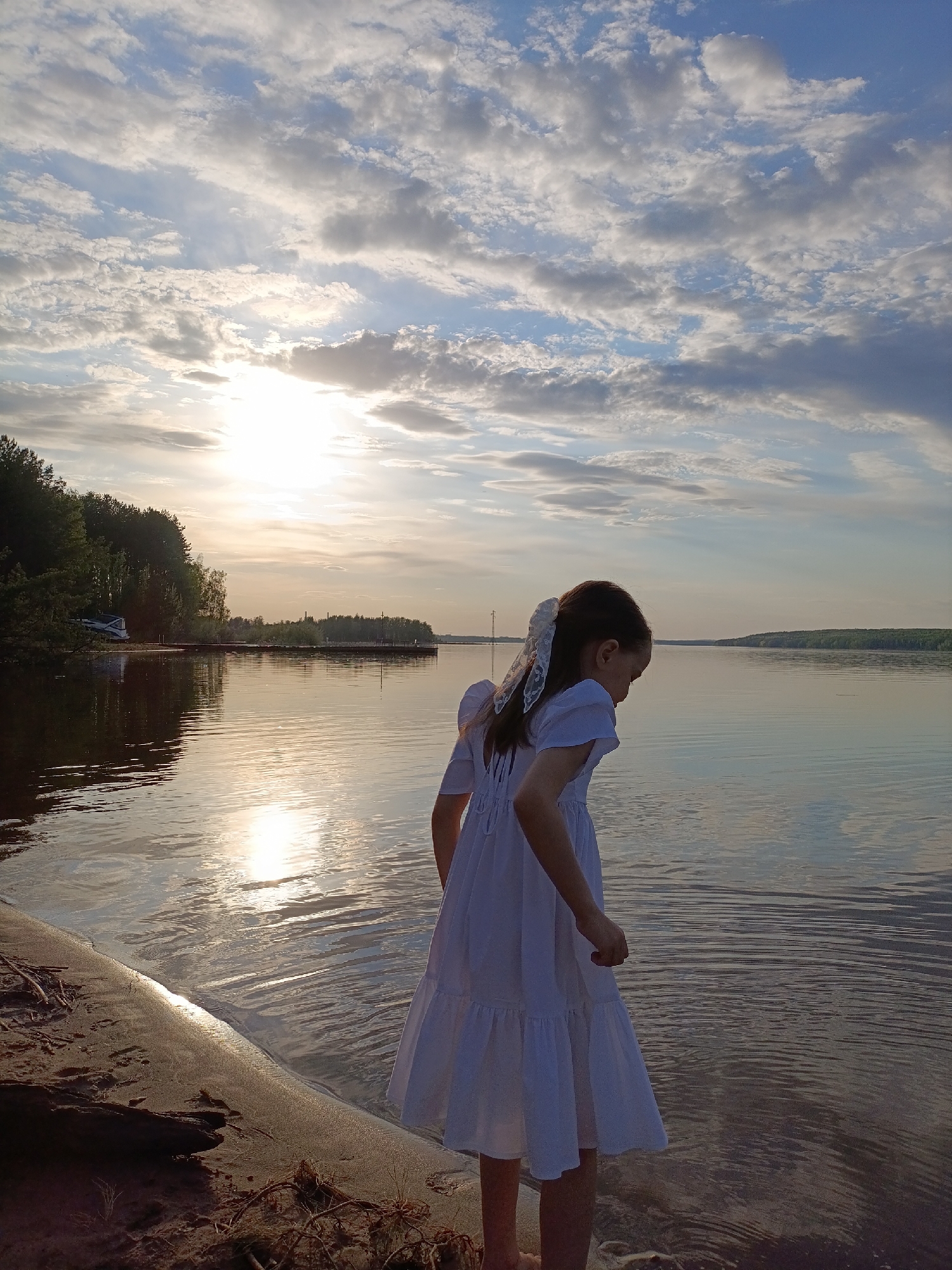 A young girl stands at the edge of a body of water. She wears a white dress and a white bow in her hair. Her feet are in the water. The setting sun reflects on the water's surface. In the background, trees line the opposite shore. The scene depicts the Chaykovsky Reservoir located in the city of Chaykovsky, Russia.