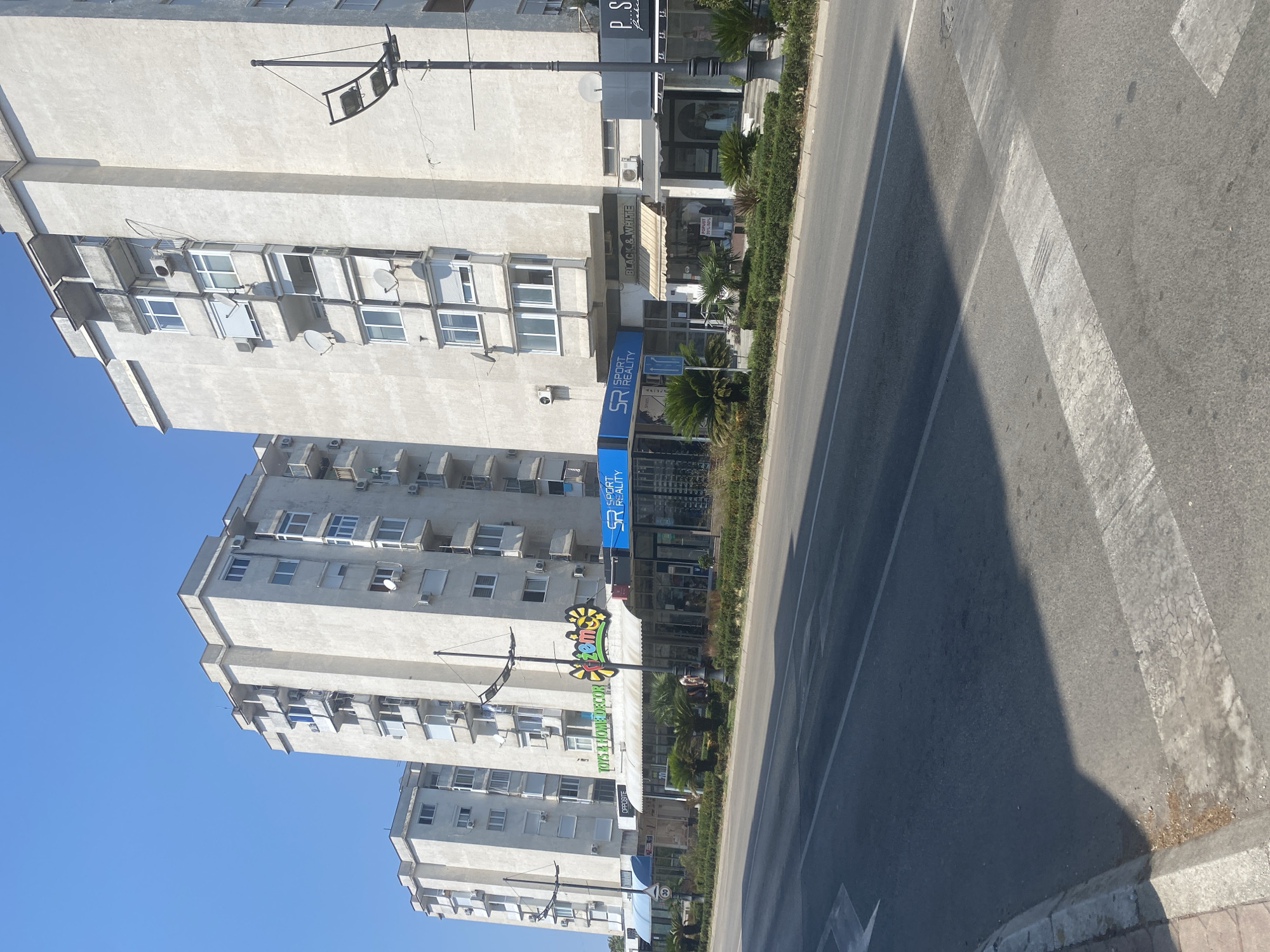 A wide shot captures a bright, sunny day in Bar, Montenegro, with the foreground dominated by an asphalt road and a pedestrian crossing. The road features white lane markings and a distinct shadow cast from an unseen object. To the right of the road, a concrete sidewalk lines the street, bordering a well-maintained green hedge.
Behind the hedge stands a row of ground-level shops with awnings, nestled at the base of three tall, white apartment buildings. These buildings, characteristic of mid-20th-century architecture, feature numerous balconies and air conditioning units. The shops display various signage, including "Toys & Home Decor," "Ftema," "Sport Reality," "Black & White," and "P.S. fashion." A few palm trees are strategically placed in front of the shops.
A single lamppost stands tall on the sidewalk, its modern design contrasting with the older buildings. The sky is a clear, deep blue, indicating fair weather. The image appears to be taken from a low angle, possibly from another vehicle on the road, suggesting a moment of transit. There are a few individuals visible near the shops, but their interactions are not discernible. The overall scene depicts a typical urban commercial street.