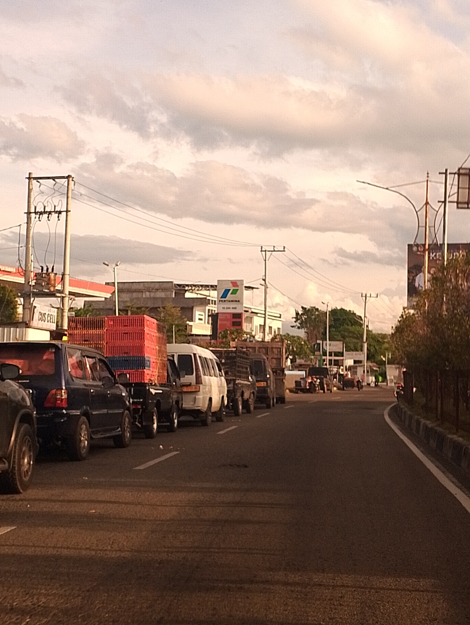A street scene in Unknown, Unknown shows a line of vehicles in traffic under a cloudy sky. The vehicles include a dark blue SUV, a dark pickup truck carrying red crates, a white van, and other vehicles further down the road. Power lines and utility poles are visible on the left side of the street, alongside buildings that appear to be commercial. A Pertamina gas station sign is also visible. The road is paved and marked with white lines. The lighting suggests it is late afternoon or early evening.