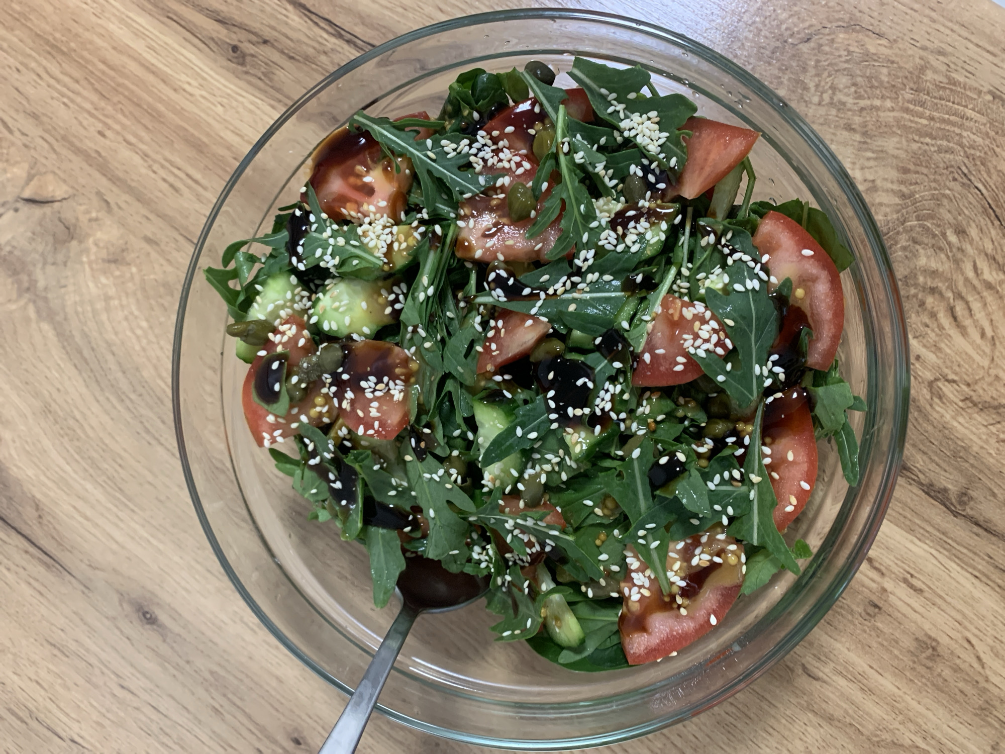 An overhead, close-up shot captures a fresh, vibrant salad served in a clear glass bowl, resting on a light brown wooden table. The salad is composed of a rich mix of ingredients, prominently featuring dark green arugula leaves, bright red quartered tomato slices, and segmented green cucumber pieces. Scattered throughout are small, dark black olives and elongated green items that appear to be capers or similar pickled vegetables. The entire dish is artfully drizzled with a dark, glossy balsamic glaze or dressing, and generously garnished with white sesame seeds, which add a contrasting texture and visual interest. A silver metal spoon is partially visible on the left side of the bowl, submerged within the salad, indicating that it is ready for consumption or in the process of being eaten. The wooden table surface features a visible grain pattern, contributing to a natural and inviting setting, likely indoors in a home environment in Ternopil, Ukraine. There is no visible text in the image.