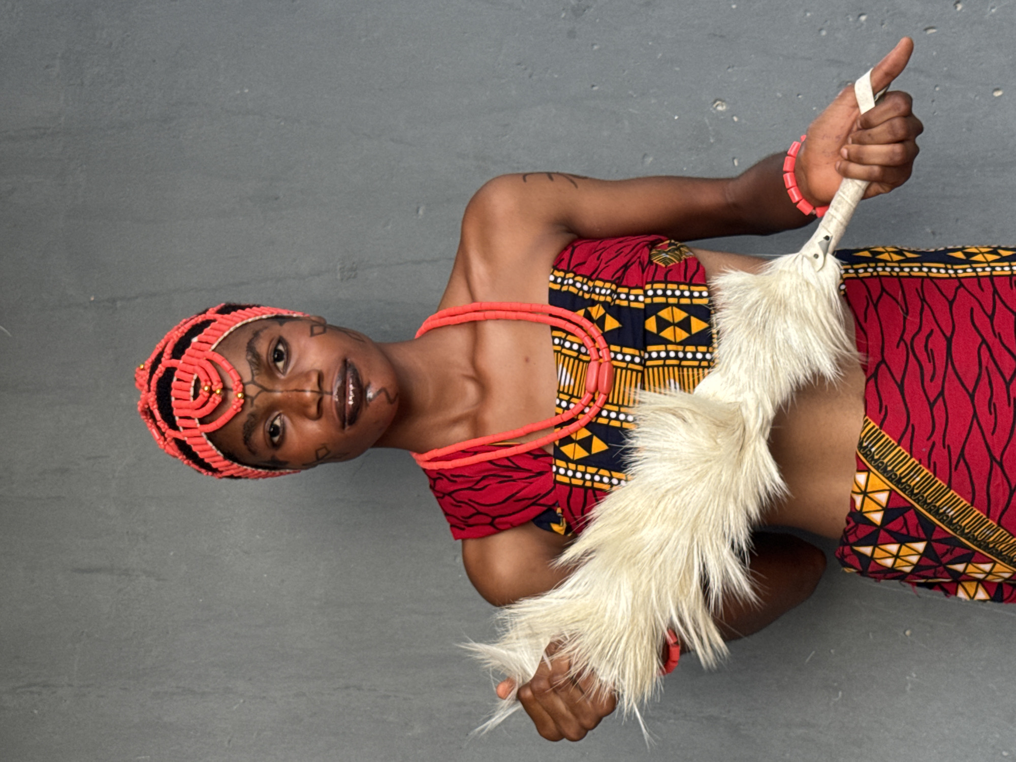A person in traditional Nigerian attire poses against a grey background. They wear a red and patterned top and skirt, a coral beaded necklace, a coral beaded bracelet, and a coral beaded headpiece. The individual holds a white, furry ceremonial item in their right hand and a tuft of the same material in their left. There is a tattoo on their left arm, and face paint marks are visible. There is no indication of a specific location in Badagry, Nigeria or any discernible location.