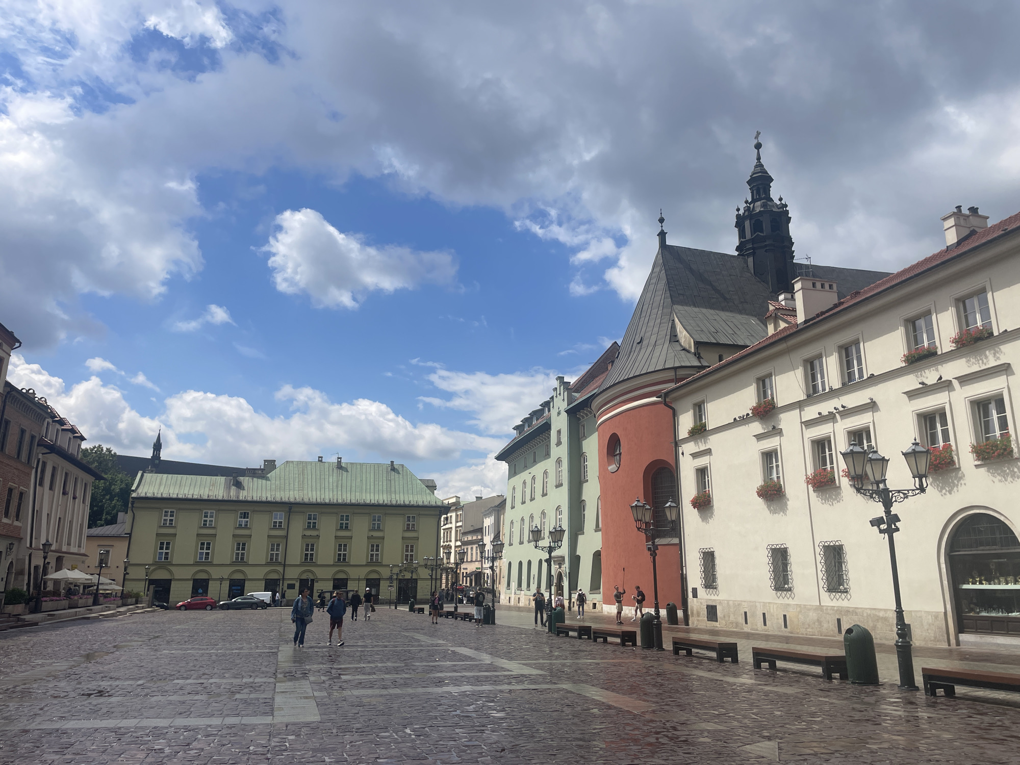 This is a wide-angle, eye-level, outdoor, daytime photograph of a European city square in Krakow, Poland, likely the Main Market Square (Rynek Główny), given the architecture. The square is paved with cobblestones, which appear wet, indicating recent rain or cleaning. A mix of historical buildings with distinct architectural styles lines the square.
In the foreground, the cobblestone pavement stretches out, with several dark wooden benches and dark green trash receptacles placed along the right side. Several lampposts with multiple lights stand along the walkway. The buildings on the right are painted in light cream colors, with windows adorned with red flowers in window boxes. One building has a striking curved facade painted in a bright coral red. Further to the right, an arched shop window displays various objects.
In the middle ground, several people are walking across the square. Two individuals, a male and a female wearing casual clothing, are walking from left to right in the center of the frame. Other individuals are visible in smaller groups or walking alone further into the square. More buildings are visible, including a large pale green building with a green roof and numerous windows. The architecture suggests a historic European city.
The background features more buildings, some with spires and pitched roofs. The sky is a vibrant blue with large, fluffy white and gray clouds scattered across it. The overall atmosphere is bright and somewhat overcast, with a sense of activity in the public space.
