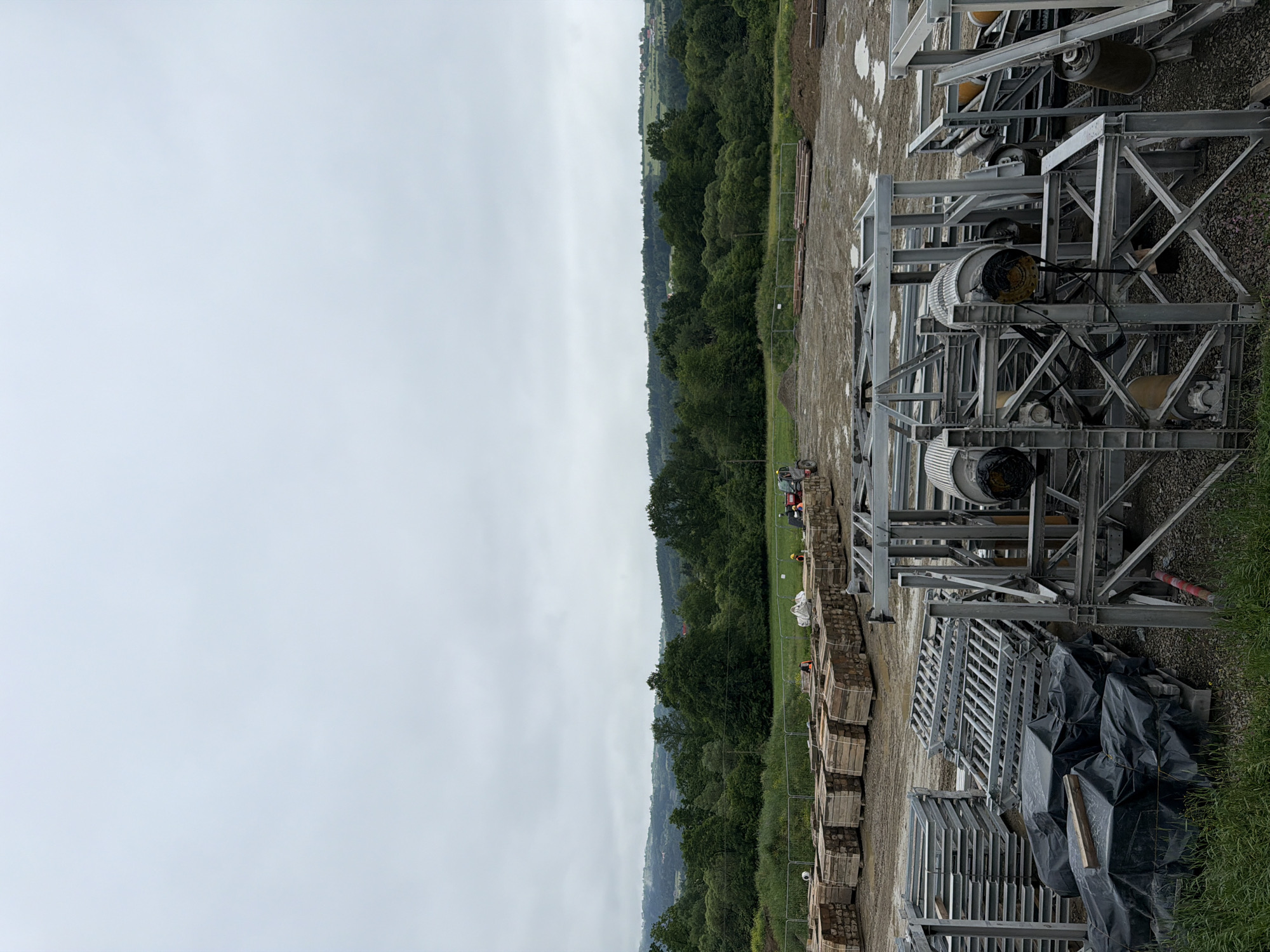 An outdoor construction site in Męcina, Poland, is depicted under an overcast sky. In the foreground, various construction materials are stacked, including wooden planks, metal scaffolding components, and items covered in black plastic sheeting. Several large, industrial metal structures with cylindrical components are also visible. In the middle ground, a field with scattered debris and a few workers can be observed. The background features a dense line of green trees and rolling hills with scattered buildings, suggesting a rural or semi-rural setting on the outskirts of a town. The overall scene conveys a sense of ongoing construction work.