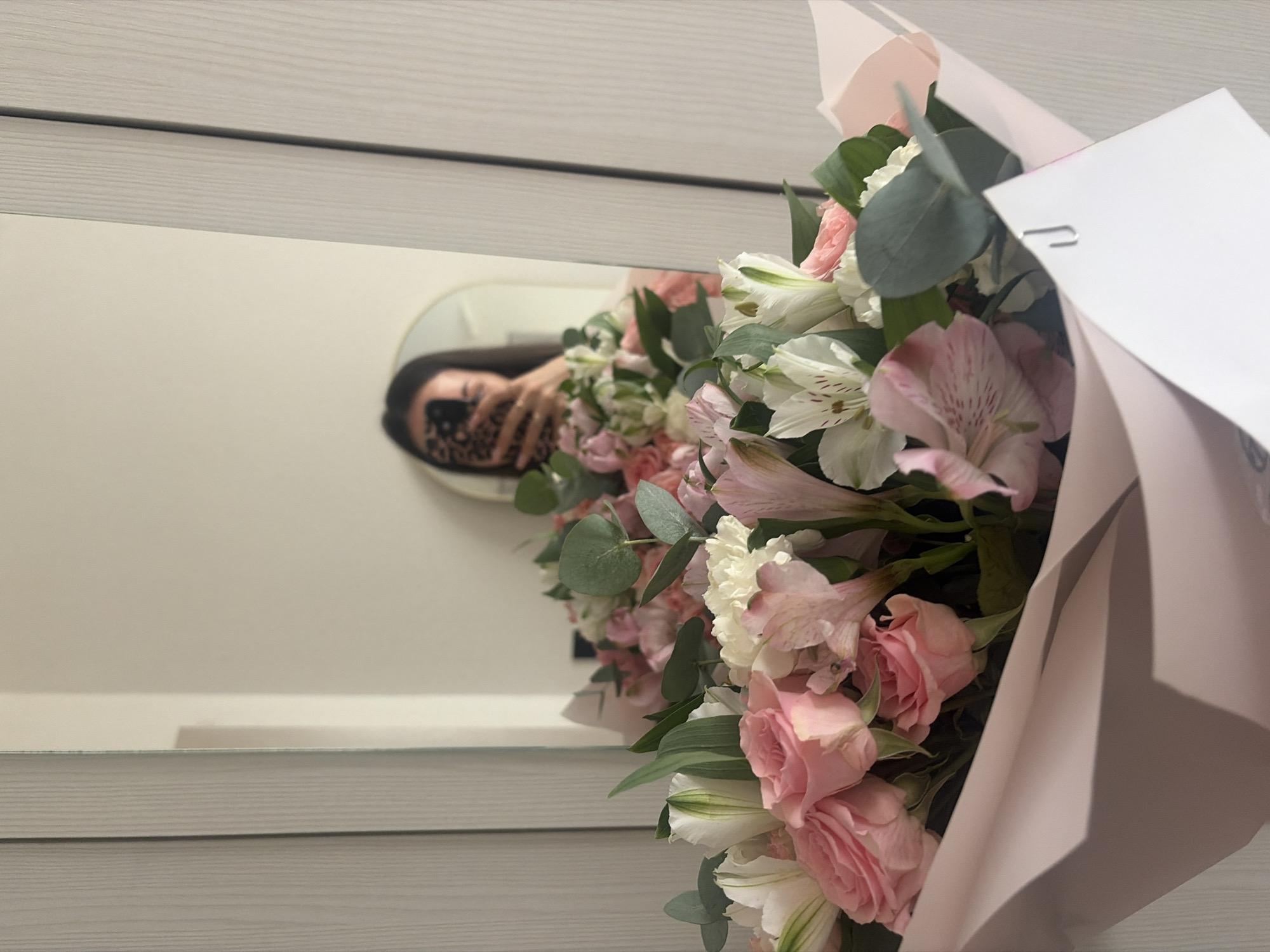 A person is taking a selfie in a mirror, holding a bouquet of pink and white flowers. The bouquet is wrapped in light pink paper. The person's face is obscured by their phone, which has a leopard print case. The background appears to be a bedroom or dressing area, with a wardrobe to the right and a mirror reflecting the scene. The overall impression is a candid moment captured while preparing to leave or documenting a gift. The scene does not provide any information that would allow for the identification of the location as Saint Petersburg, Russia.