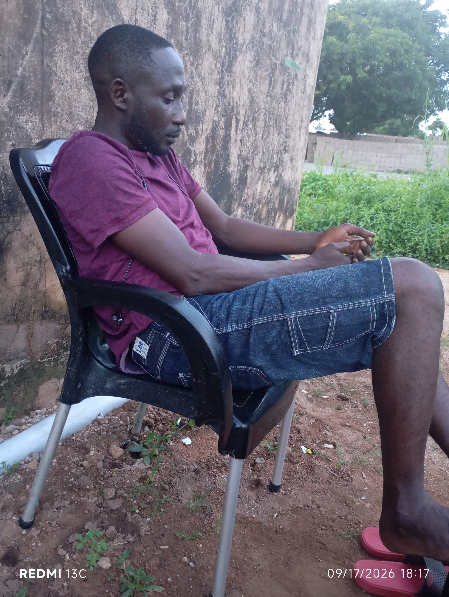 An individual, appearing to be a Black male, is seated outdoors on a black plastic chair. He is wearing a maroon t-shirt and denim shorts. His hands are clasped together in his lap, and he is looking down. The chair is positioned on uneven, reddish-brown soil with scattered rocks and sparse green vegetation. In the background, a textured, light-colored wall is visible, along with a dense green bush and trees. A partially visible brick wall and some stacked construction materials are also present in the distance. The overall setting suggests a rural or semi-urban environment, possibly in Shendam, Nigeria, given the provided context, though the image itself does not contain explicit geographical markers for this specific location. The lighting indicates it is daytime. A timestamp and device model, "REDMI 13C 09/17/2026 18:17", are superimposed on the image.