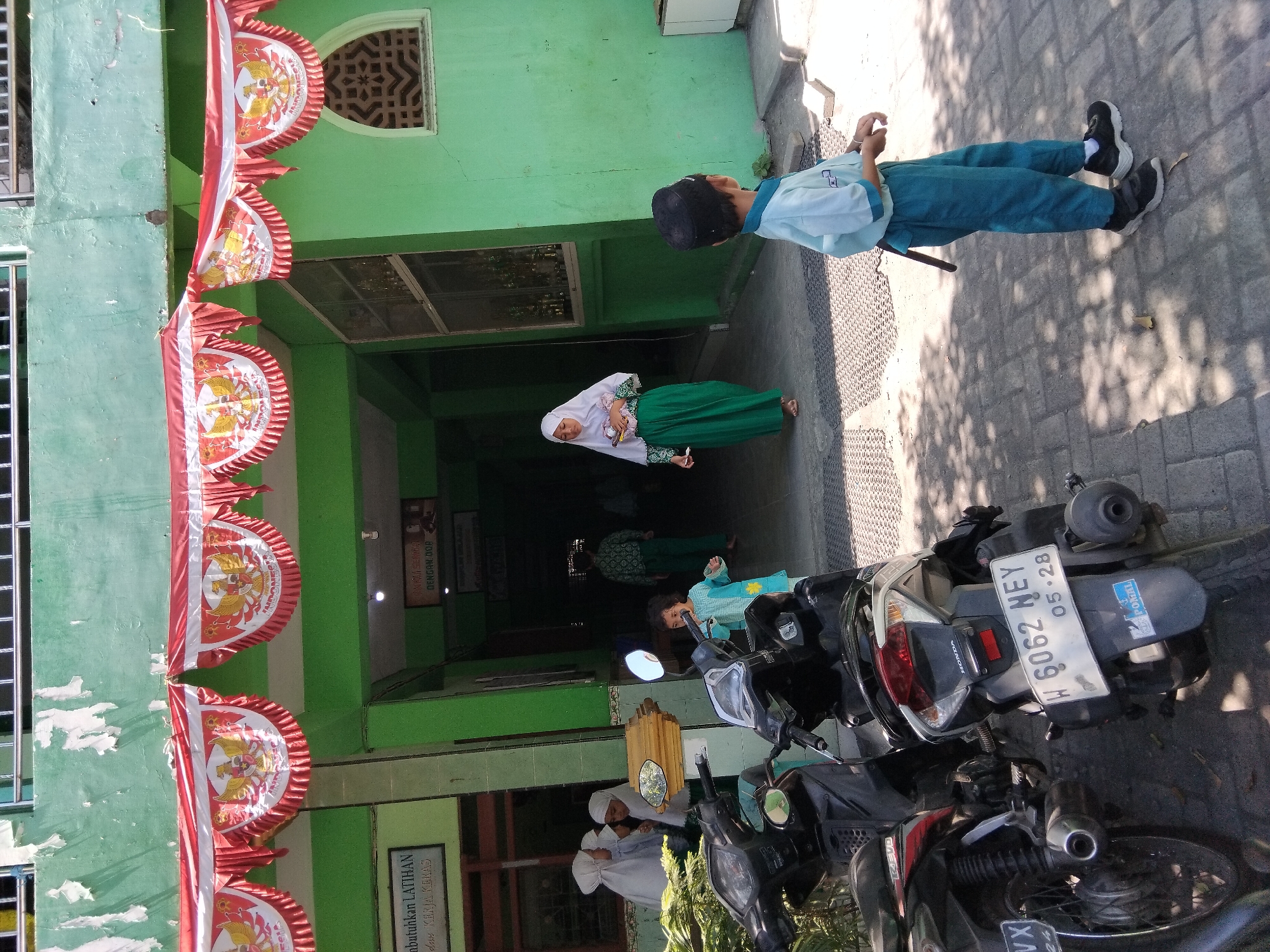 The image depicts a scene outside a building with green walls, likely a school, in Kepuh Kiriman, Indonesia. Red and white decorative banners with the Garuda Pancasila symbol and the word "INDONESIA" are hung across the upper facade. Several motorbikes are parked in the foreground. In the middle ground, a young child wearing a blue tunic and pants stands near a motorbike, appearing to be playing or looking around. Further back, a girl in a white hijab and green skirt is walking towards the building's entrance. To the left, a group of children in white hijabs are gathered near a doorway. A boy wearing a light blue uniform shirt and dark pants, with a black skullcap, is standing in the foreground, facing away from the camera. The ground is paved with textured tiles and some areas have dappled sunlight. The building has arched windows and a framed sign with text is visible on the wall to the left.