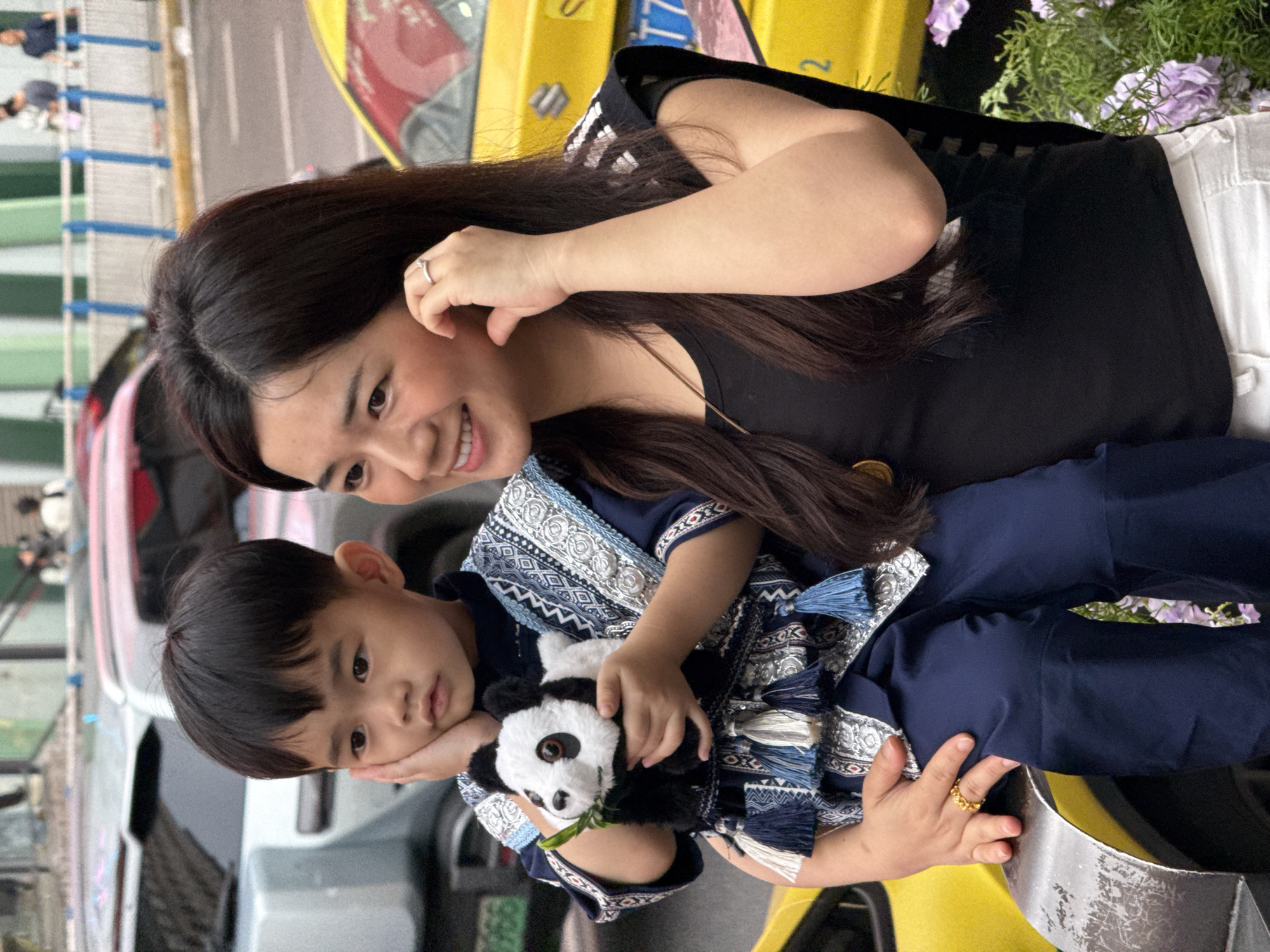 In Liangjiang, China, a woman and a child are situated near a yellow vehicle. The woman has long, dark hair and wears a black top. She is smiling and has a ring on her finger. The child is wearing a blue and white patterned outfit and holding a panda toy. The child's hand is resting on his face. In the background, there are other vehicles and people. There are also some plants with purple flowers.