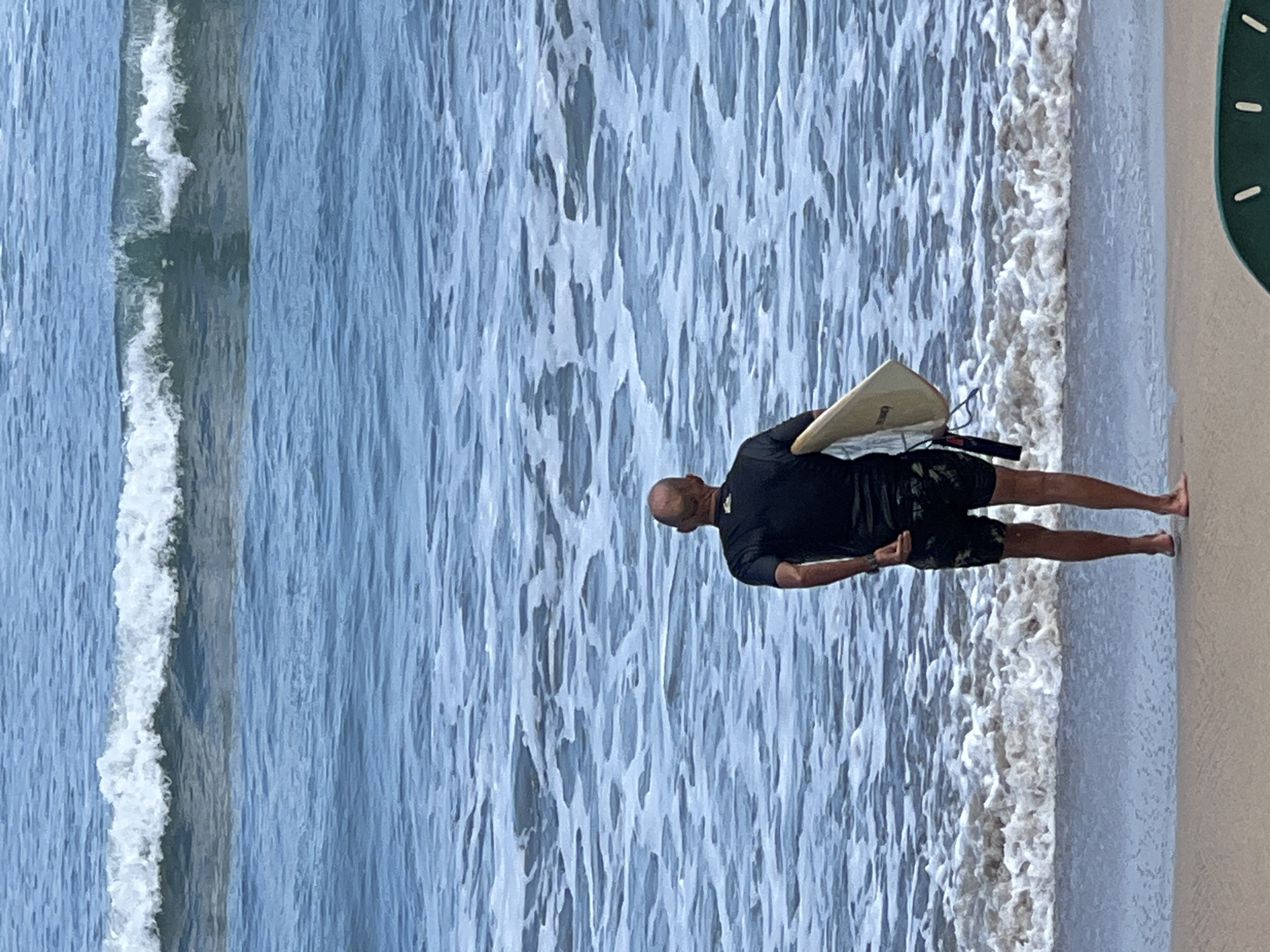 A lone, bald man wearing a black rashguard and camouflage shorts stands on the sandy shore of Kuta Beach, Indonesia. He is facing the ocean, holding a light-colored surfboard under his right arm. The ocean is a vibrant blue, with several waves breaking in the distance, creating white foam that extends towards the shore. The sky is not visible in the frame. The image captures a moment of quiet anticipation as the surfer prepares to enter the water.