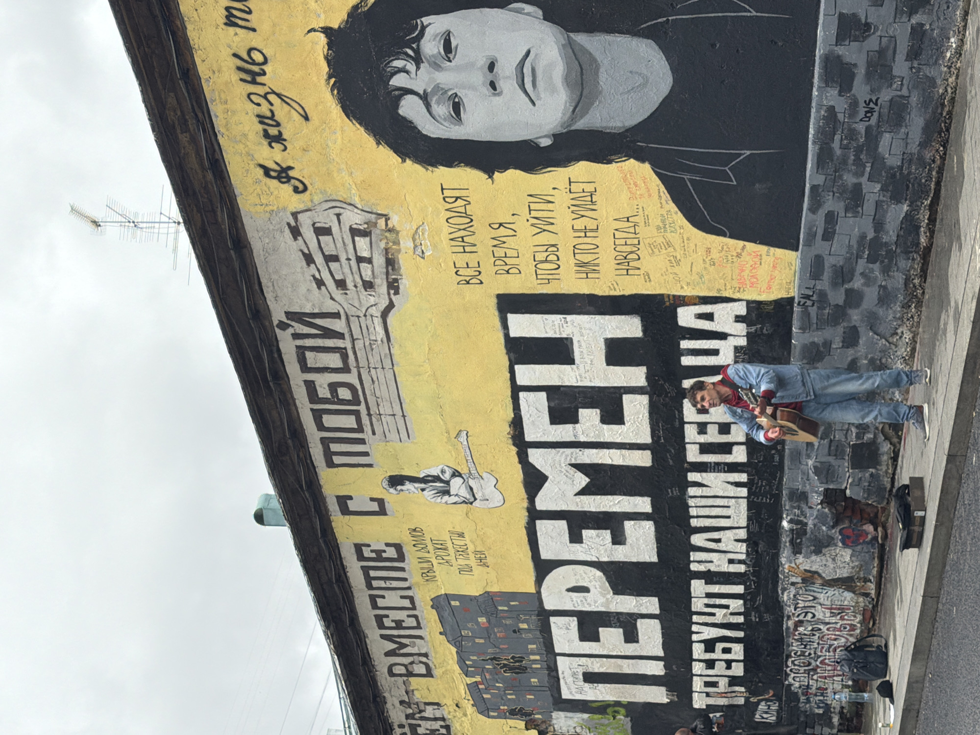 This is a vertical, eye-level, outdoor shot of a mural on a building. The mural features a large black and white portrait of a young man with dark, messy hair and solemn eyes. To the left of the portrait, large, stylized black lettering on a yellow background reads "ЕМ ВМЕСТЕ С ТОБОЙ" (Together with you). Below this, there is a depiction of a building and a smaller image of a man playing a guitar. To the right of the portrait, more black lettering on a yellow background says "ВСЕ НАХОДЯТ ВРЕМЯ, ЧТОБЫ УЙТИ, НИКТО НЕ УЙДЕТ НАВСЕГДА..." (Everyone finds time to leave, no one will leave forever...). Below this, on a black background, are the words "ПЕРЕМЕН ТРЕБУЮТ НАШИ СЕГАЦА" (We demand change now).
In the lower right portion of the image, a young man dressed in a blue denim jacket, a red shirt, and blue jeans is standing on a sidewalk and playing an acoustic guitar. A black guitar case lies on the ground near him, suggesting he may be a street performer. The overall scene appears to be an urban environment with the mural dominating the wall. The sky is overcast, indicating potentially cloudy or dreary weather. The mural and the presence of graffiti suggest a location with a vibrant street art culture.