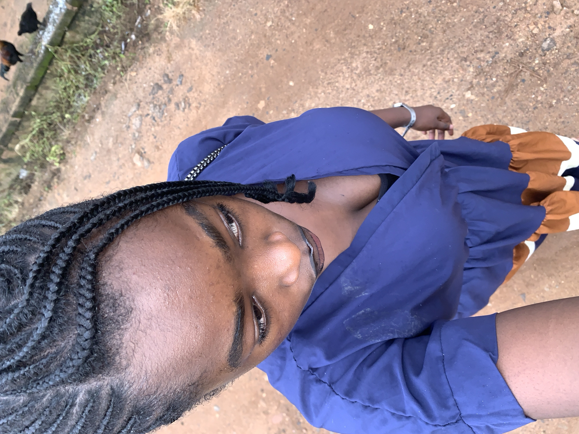 A young Black woman with braided hair and dark skin is depicted from a high-angle shot. She is wearing a dark blue dress with ruffled details and a white and orange striped hem. A silver watch is visible on her left wrist. The background appears to be an outdoor, dusty environment with a dirt path and some vegetation. In the upper right corner, two chickens are visible, one dark and one with red and black plumage. The setting is identified as Rafin Par, Nigeria.