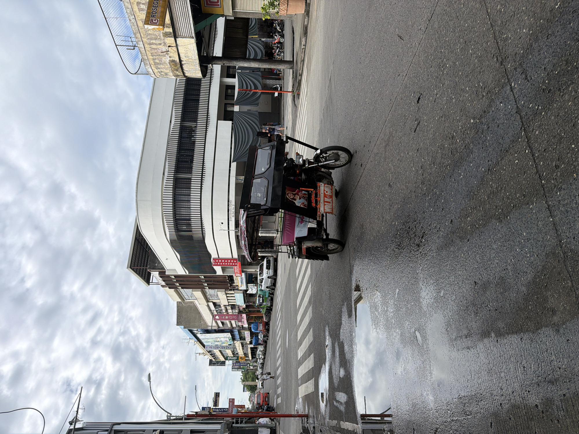 The image captures a street scene in Davao City, Philippines. A motorized tricycle (tuk-tuk) is parked on the right side of the road near a crosswalk. The street is wet, reflecting the cloudy sky and surrounding buildings. Several buildings line the street, with signs visible in a mixture of English and possibly Tagalog. Other vehicles are parked further down the street. There are no people visible immediately near the tricycle, but there might be someone in the driver's seat.