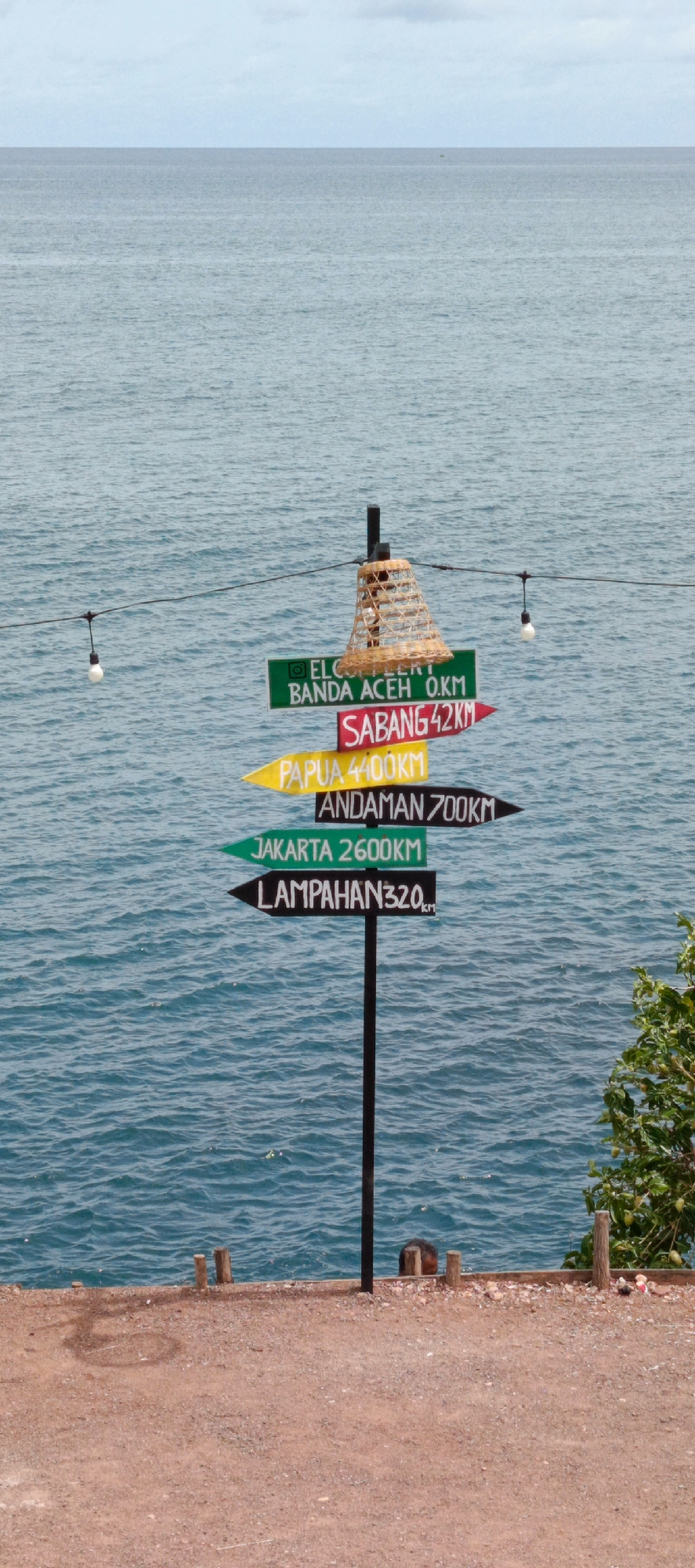 An outdoor signpost displays distances to various locations from what appears to be a coastal viewpoint. The signpost is black and has several directional arrows. The top arrow, in green, reads "ELCG. / BANDA ACEH 0.KM". Below that, a red arrow points to "SABANG 42KM". A yellow arrow indicates "PAPUA 4400KM", followed by a black arrow pointing to "ANDAMAN 700KM". The next green arrow points to "JAKARTA 2600KM", and the bottom black arrow reads "LAMPAHAN 320KM". A woven lamp shade hangs from a wire strung across the scene, with a single light bulb attached below it. The signpost stands on a reddish-brown, gravelly ground, with wooden posts in the foreground. The background is dominated by a calm, blue ocean under a pale blue sky. A small portion of green foliage is visible on the right side of the image. The signpost is located in Banda Aceh, Indonesia, as indicated by the inscription on the topmost sign.