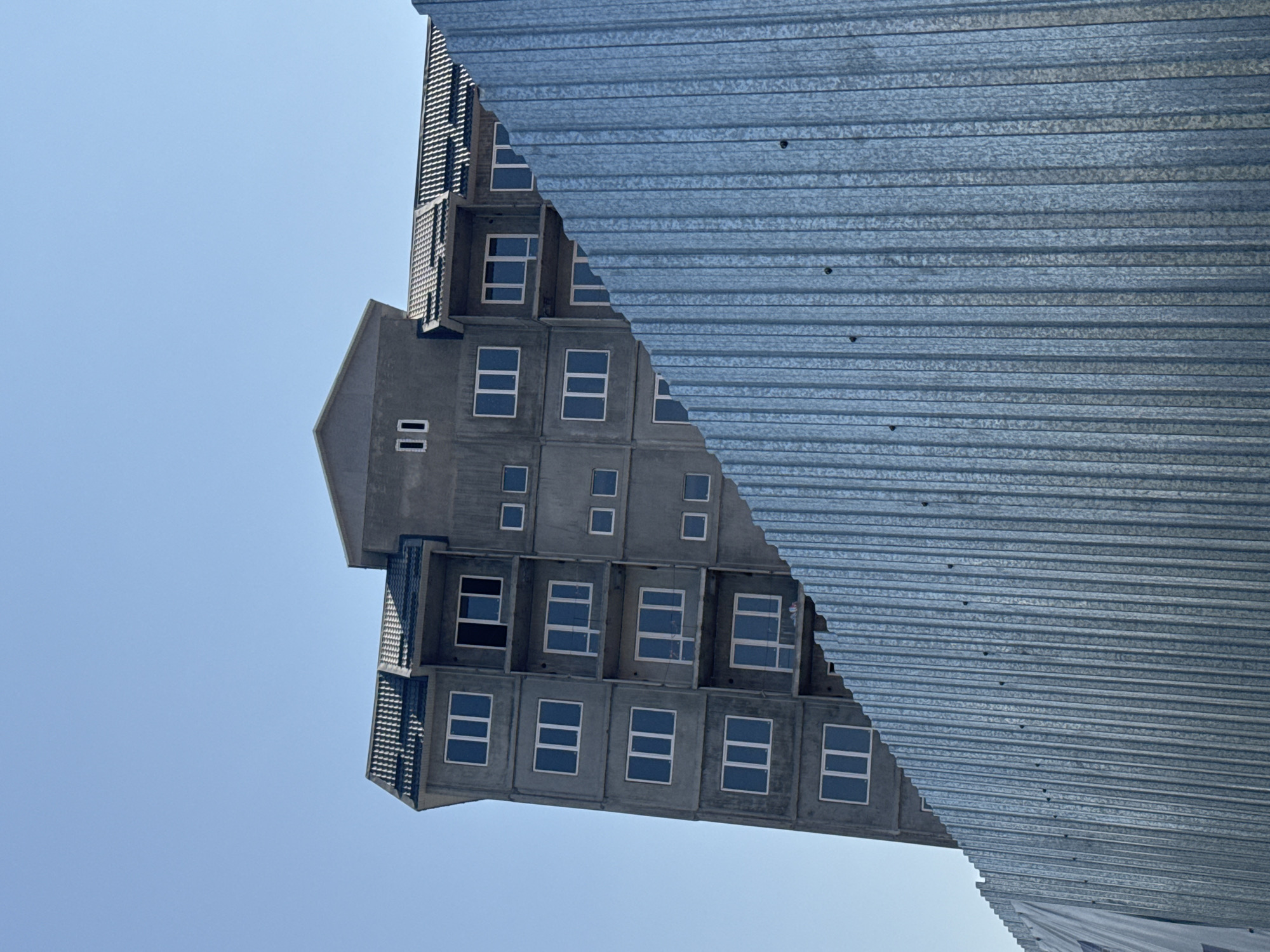 The image depicts a tall, unfinished concrete building against a clear blue sky. The building has multiple floors with numerous windows, most of which are glassed in. A corrugated metal fence runs across the foreground, partially obscuring the lower portion of the building. The fence appears to be a temporary barrier for a construction site. There are no people or vehicles visible in the scene. The overall impression is of urban construction in progress. The image is taken from a low angle, looking up at the building.