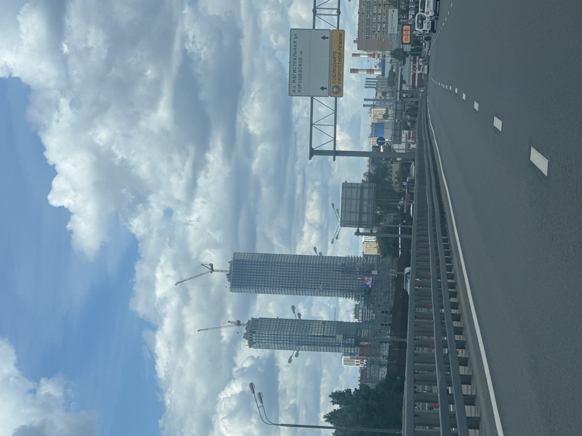 The image shows a view from a car on a highway in Moscow, Russia, with two tall skyscrapers under construction in the background. The skyscrapers are made of glass and steel, with cranes on top. The sky is cloudy, with patches of blue visible. There is a traffic sign indicating "4-ya Magistralnaya Ul., Khoroshevskoye Sh." and a warning to observe speed limits. In the distance, industrial buildings and chimneys are visible. The highway has multiple lanes and a guardrail on the left side. Several cars are on the road.