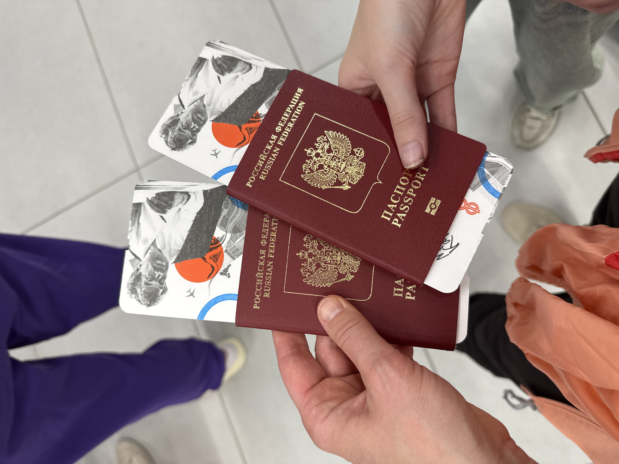 A person is holding two Russian passports and two boarding passes in their hands. The passports are maroon-colored with gold lettering and the Russian coat of arms. The boarding passes have a black and white image of a man's face with a red circle and an airplane icon on them. The person is standing on a tiled floor. There are other people in the background with various clothing and shoes. Based on the tile and color scheme, it is impossible to confirm it is Ufa, Russia.
