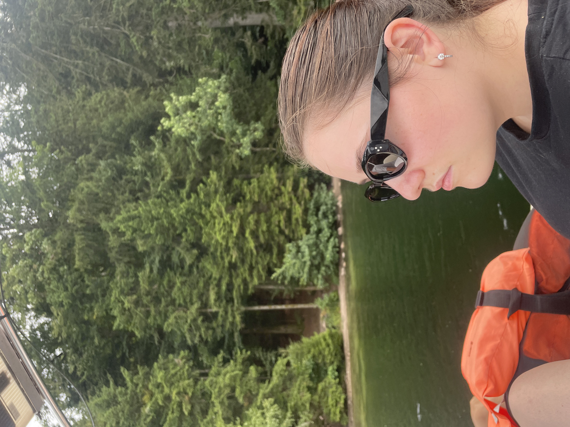 A young woman wearing black sunglasses and a black t-shirt is on a boat. She is wearing a diamond stud earring. In the background, there is a body of water and a dense forest with tall green trees. The water appears calm and dark green, reflecting the surrounding foliage. The boat has an orange life jacket visible in the foreground. The image is taken from a medium close-up perspective, focusing on the woman's profile. The scene suggests a peaceful outing on a lake or river surrounded by nature. The location is not identifiable as Gérardmer, France, based on the visual information provided.